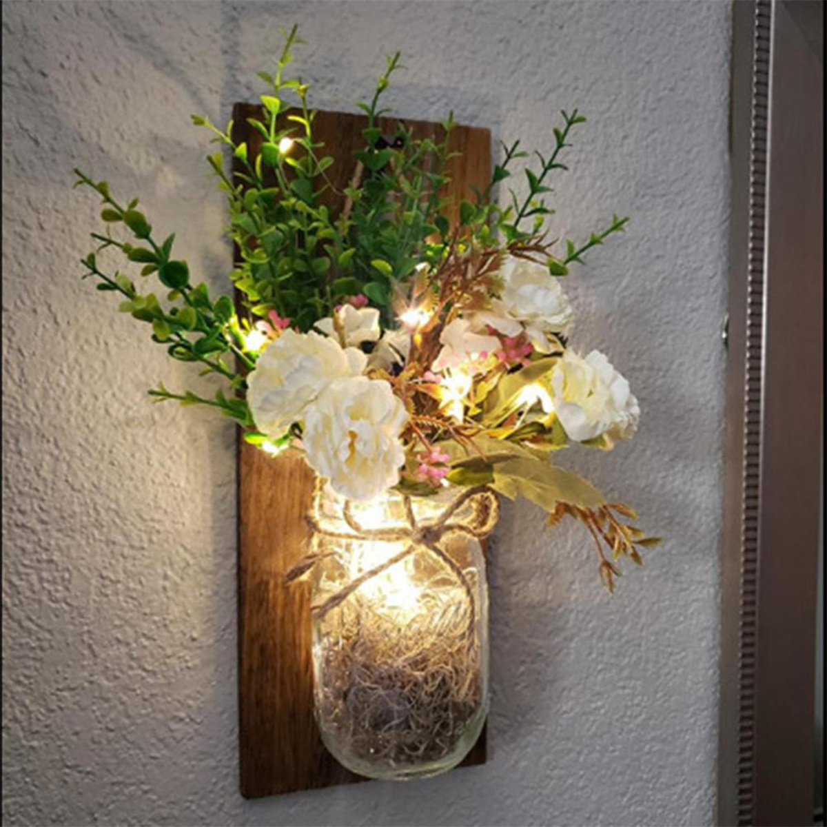 Eine Wanddekoration mit Blumen in einem Glas, beleuchtet von Lichterketten, auf einer Holzplatte.