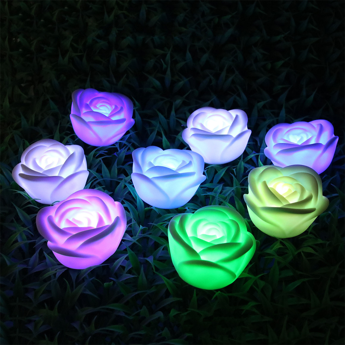 Sieben leuchtende rosenförmige Lichter in verschiedenen Farben auf grünem Gras.