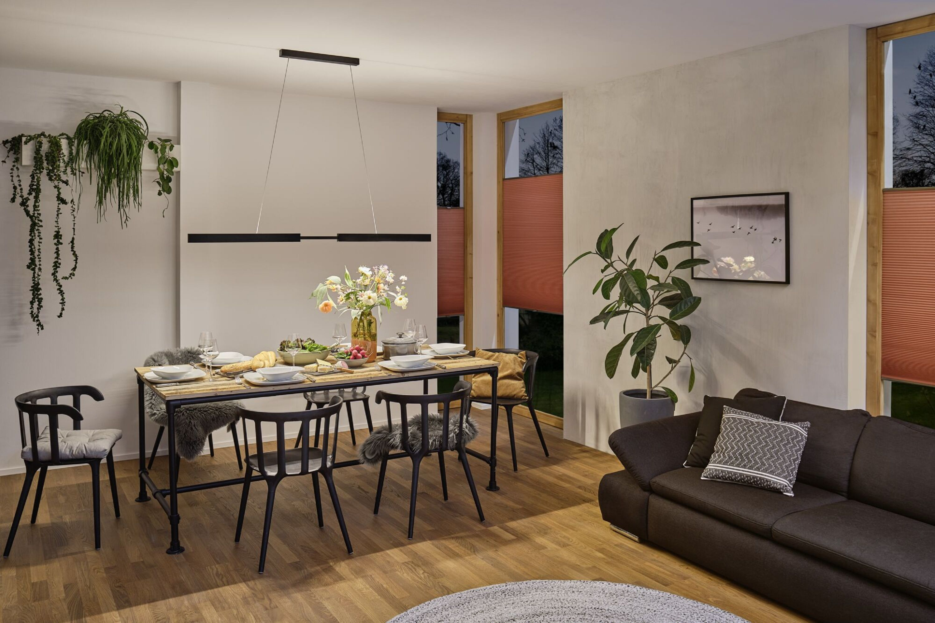 Jadalnia z długim stołem, krzesłami i sofą. Widoczne są rośliny i okna.