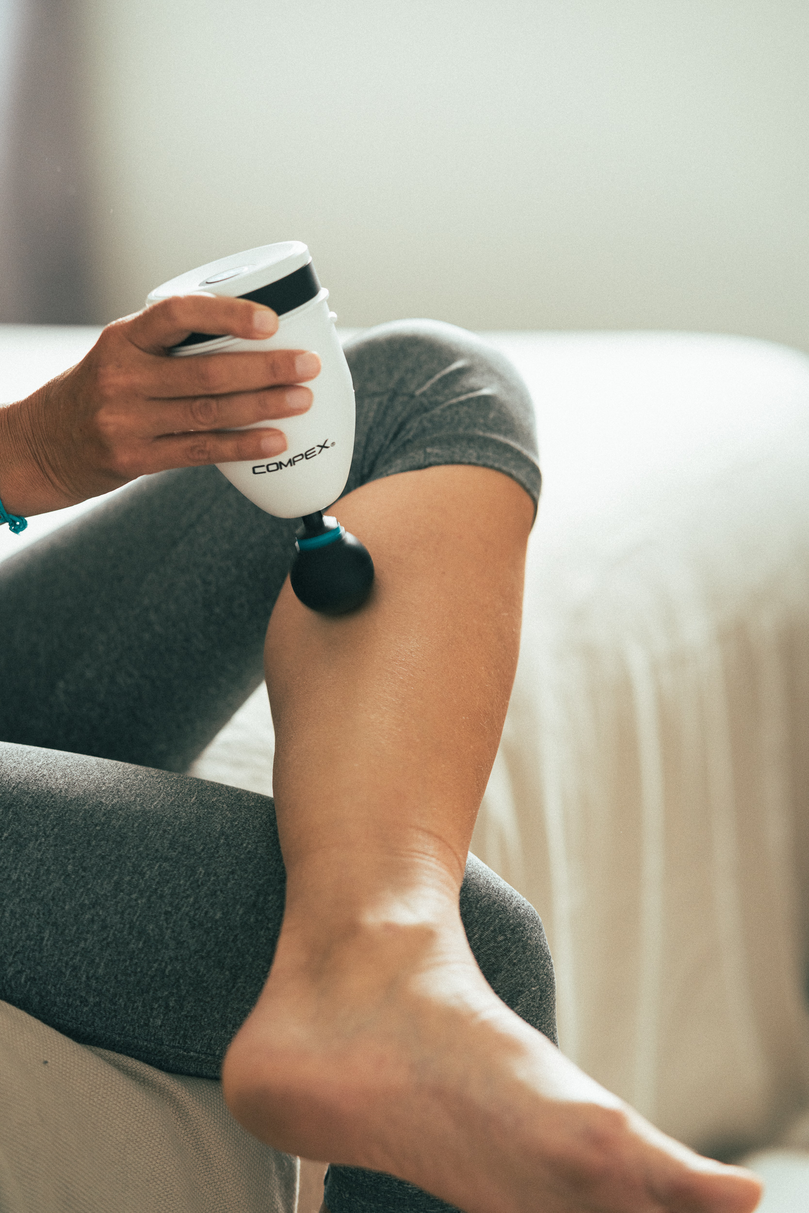 Eine Person massiert ihr Bein mit einem weißen und schwarzen Compex-Massagegerät. Sie trägt graue Leggings. Ein weißes Bett im Hintergrund.