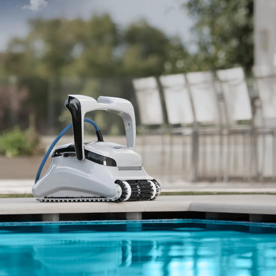 Weißer Roboter-Poolreiniger am Poolrand mit blauem Wasser. Stühle im Hintergrund.