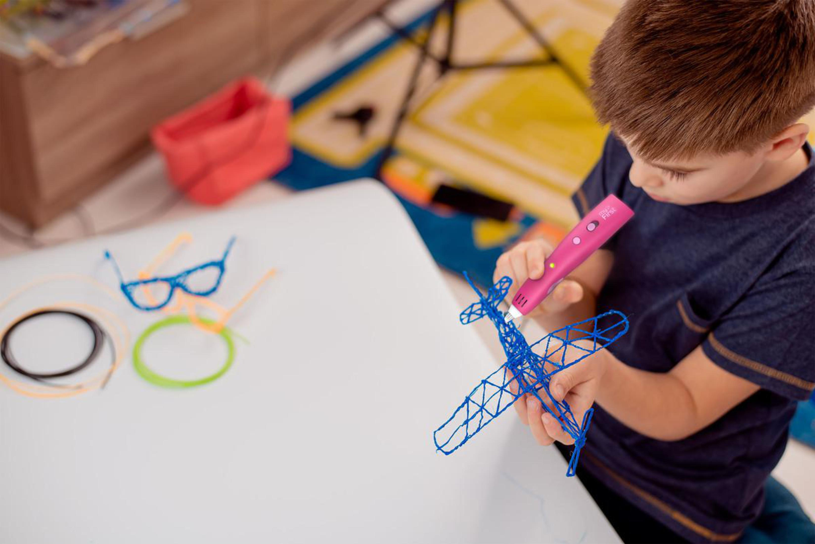 Ein Junge benutzt einen pinkfarbenen 3D-Stift, um ein blaues Flugzeug zu bauen. Andere 3D-Designs auf dem Tisch.