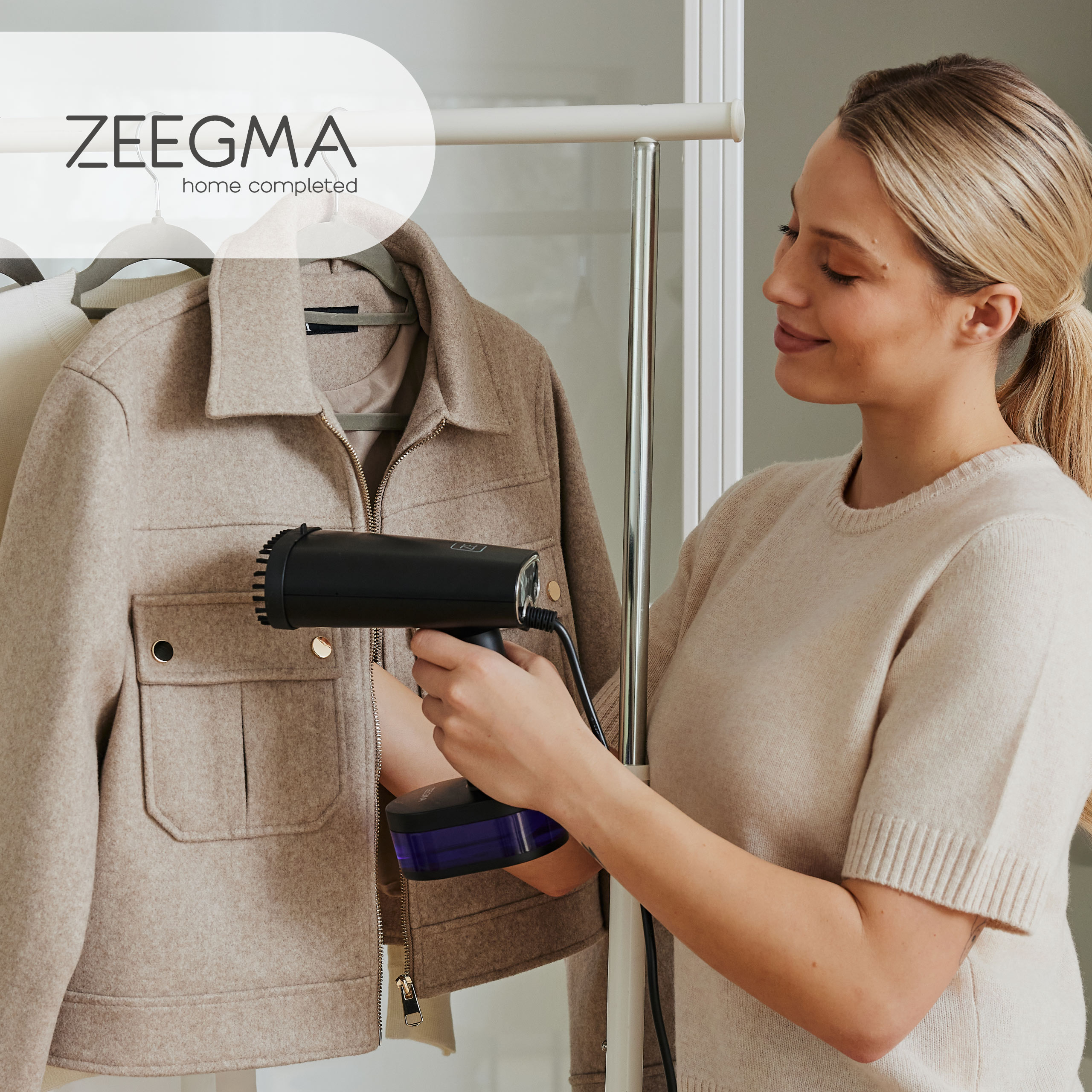 Kobieta paruje kurtkę. Czarny parownik w dłoni, beżowa kurtka na wieszaku, logo.