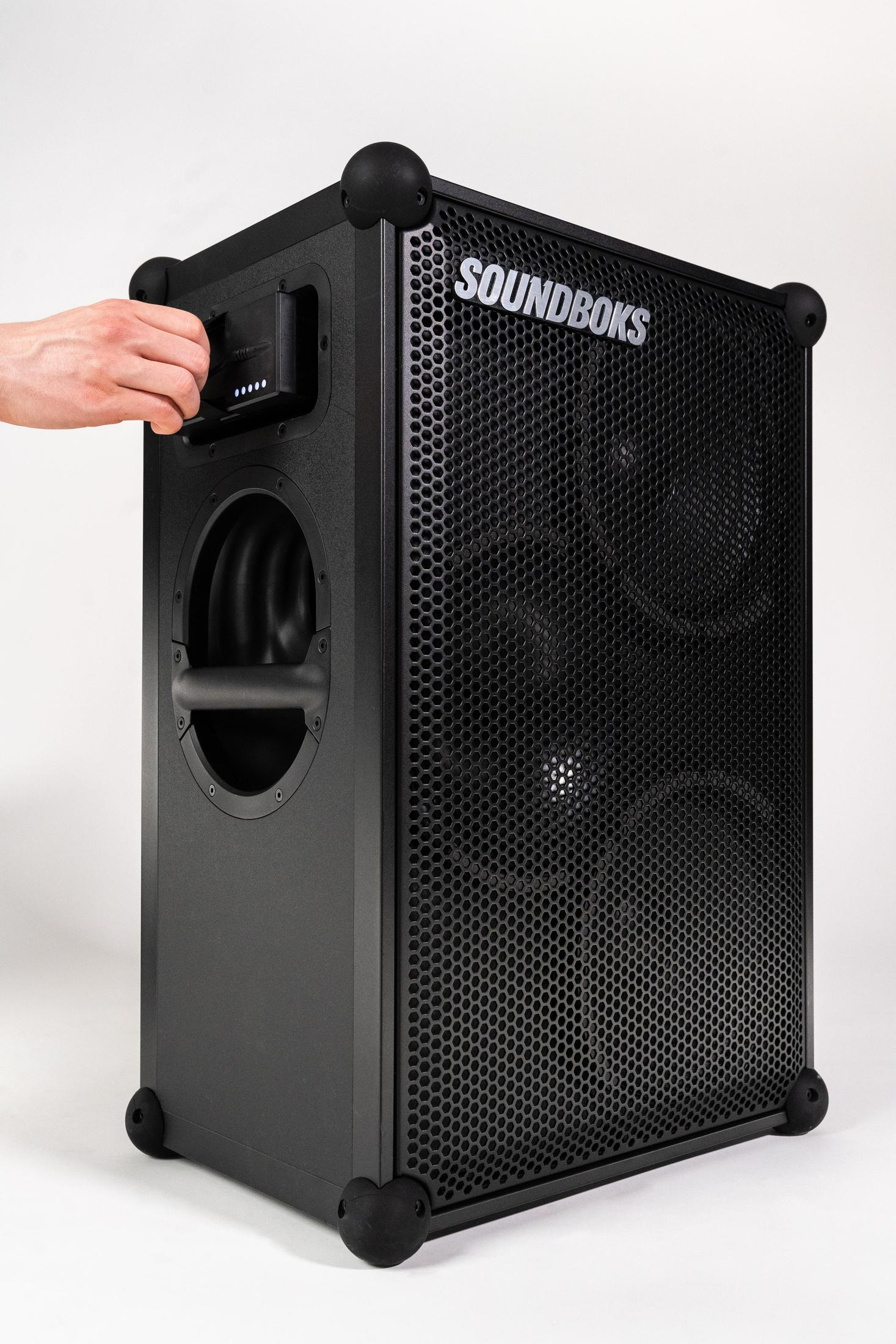 Schwarzer SOUNDBOKS-Lautsprecher, eine Hand berührt das Bedienfeld, vor weißem Hintergrund.
