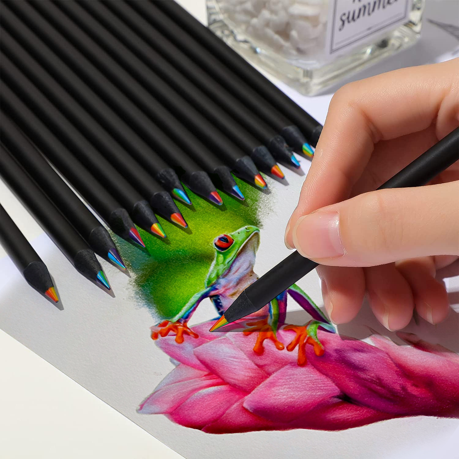 Mano dibujando una rana con un lápiz. Lápices y un recipiente transparente en el fondo.