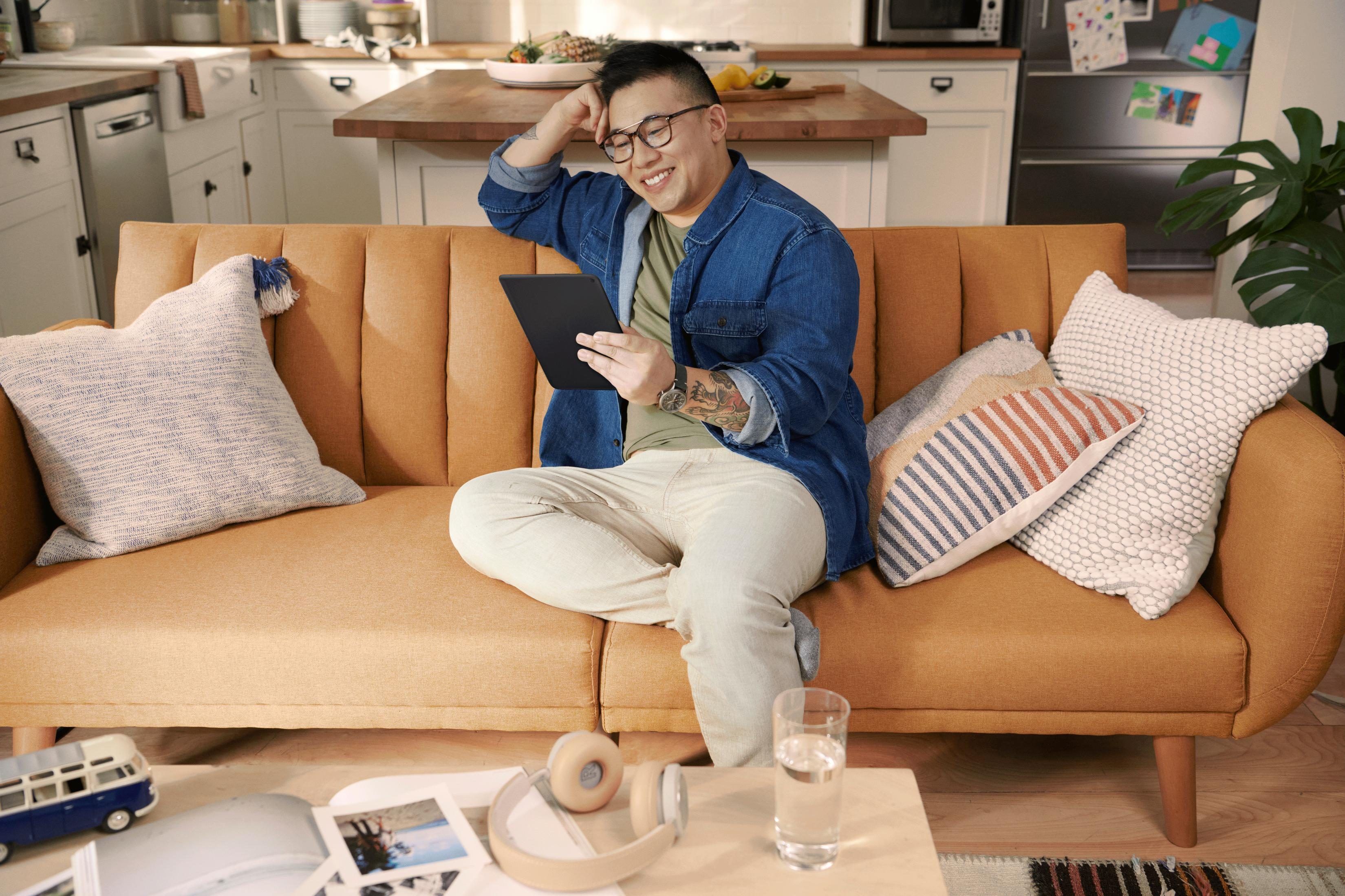 Ein Mann in Jeansjacke sitzt auf Sofa, hält Tablet, lächelt.