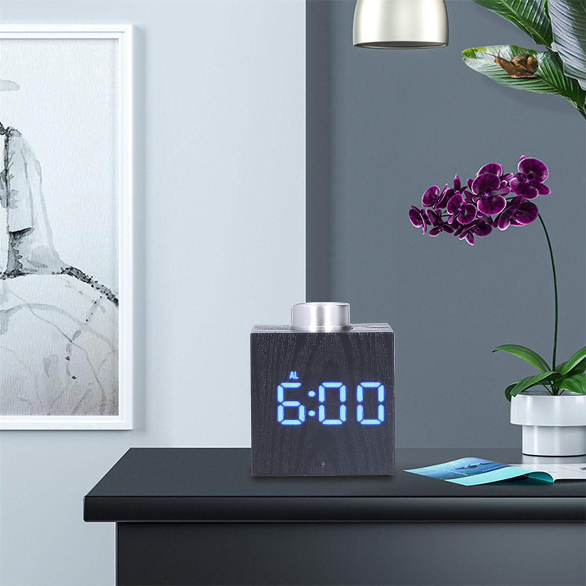 Schwarze Holzwürfeluhr mit blauem LED-Display, das 6:00 anzeigt. Schwarzer Tisch, weiße Wand, Pflanze.