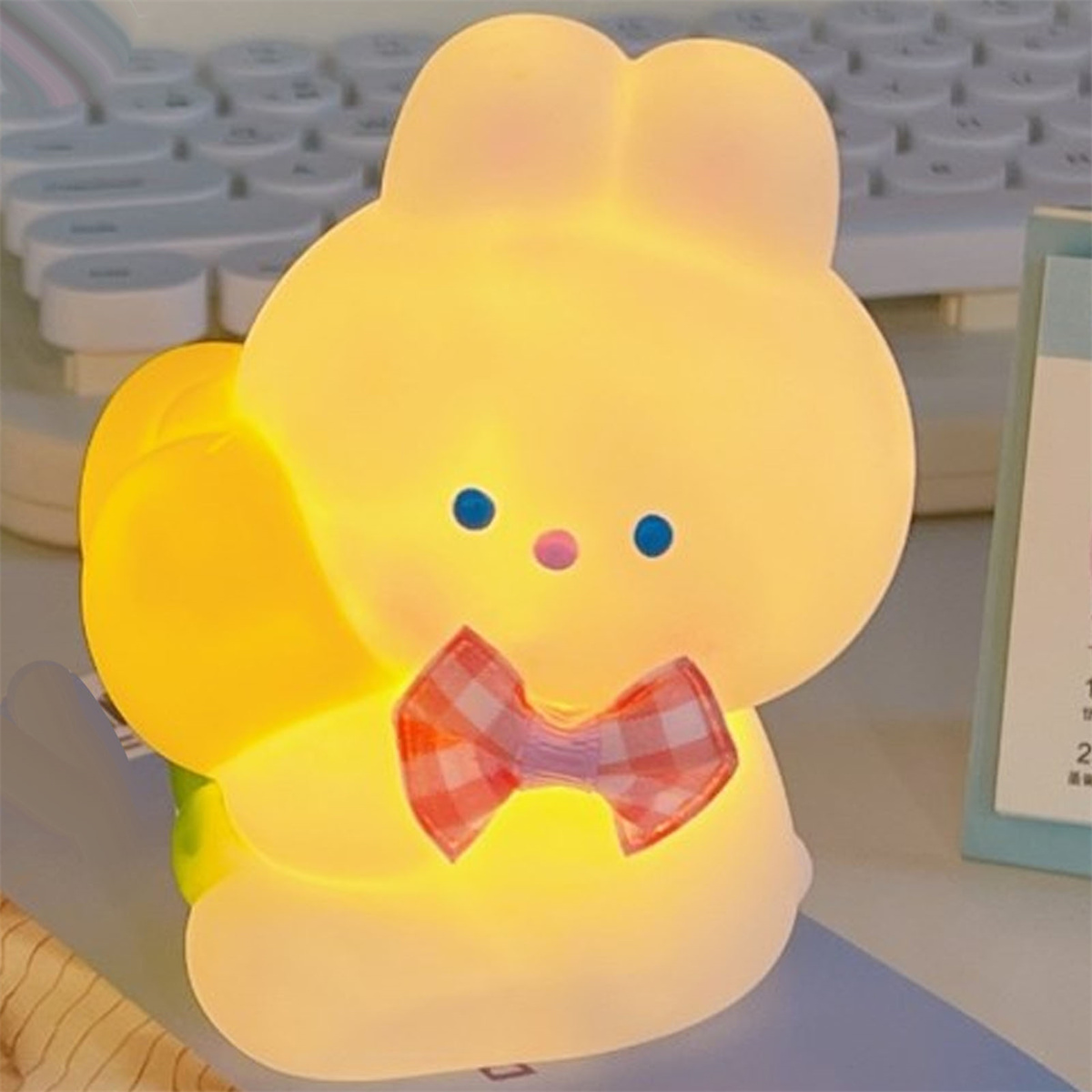 Eine leuchtende, gelbe hasenförmige Lampe mit roter karierter Schleife, die auf einem Schreibtisch steht.
