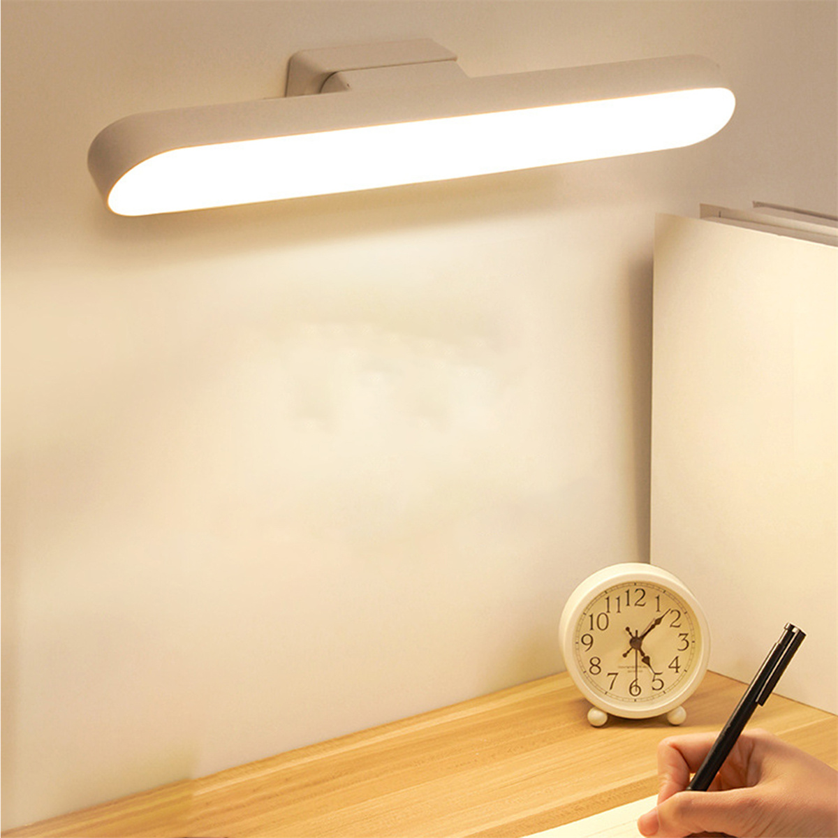 Eine Schreibtischlampe leuchtet auf einem Schreibtisch mit Uhr, Stift und Notizblock. Weiße Wand im Hintergrund.