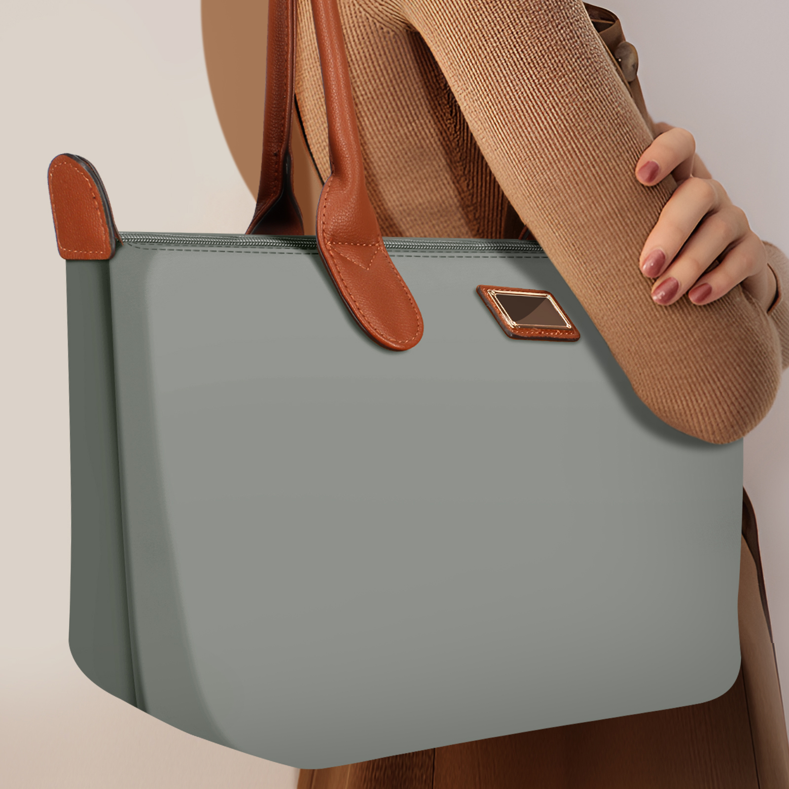 Bolso tote gris con asas marrones sostenido por una persona. Suéter beige. El bolso tiene una etiqueta dorada rectangular.