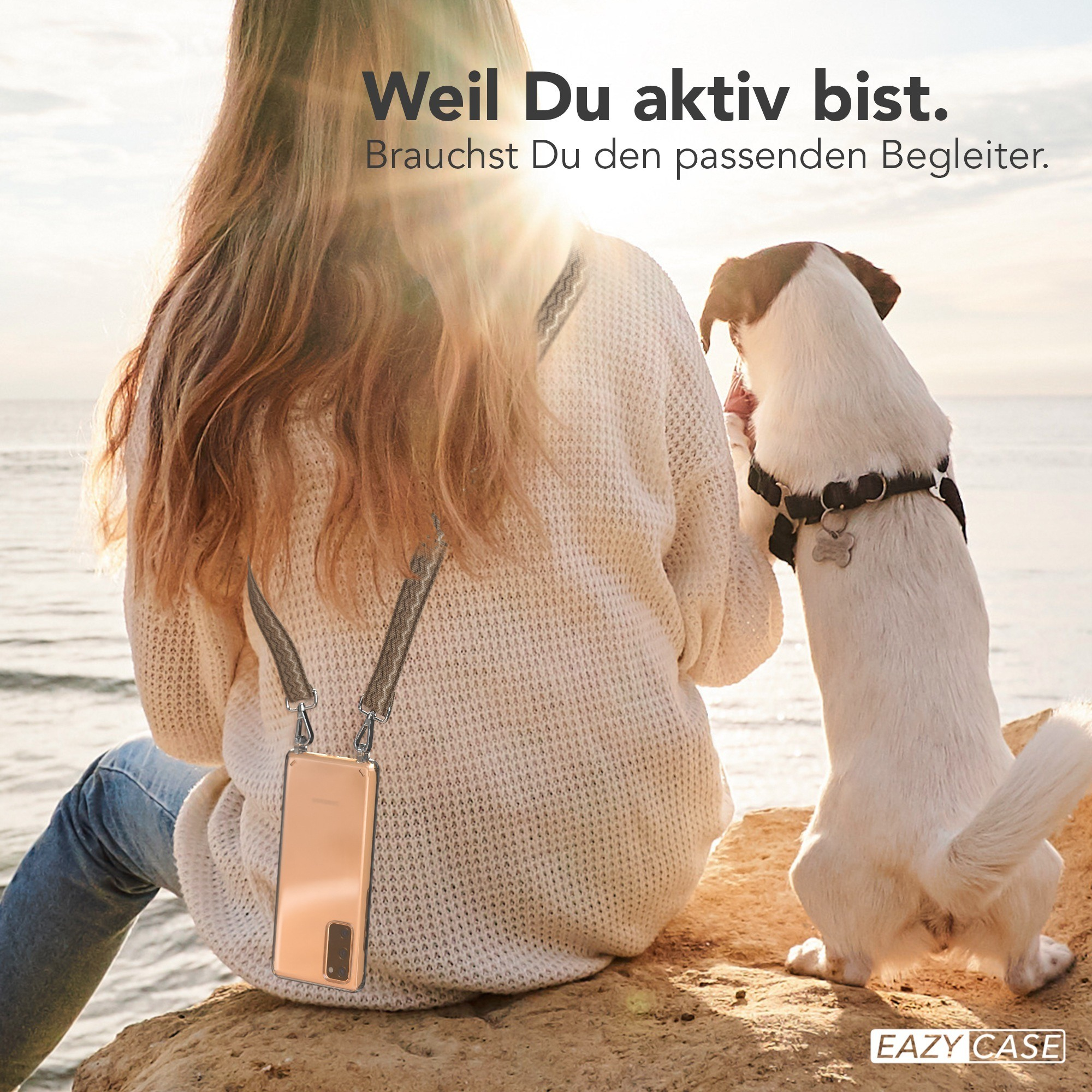 Eine Frau mit Handyhülle und Hund am Meer, die Sonne scheint.