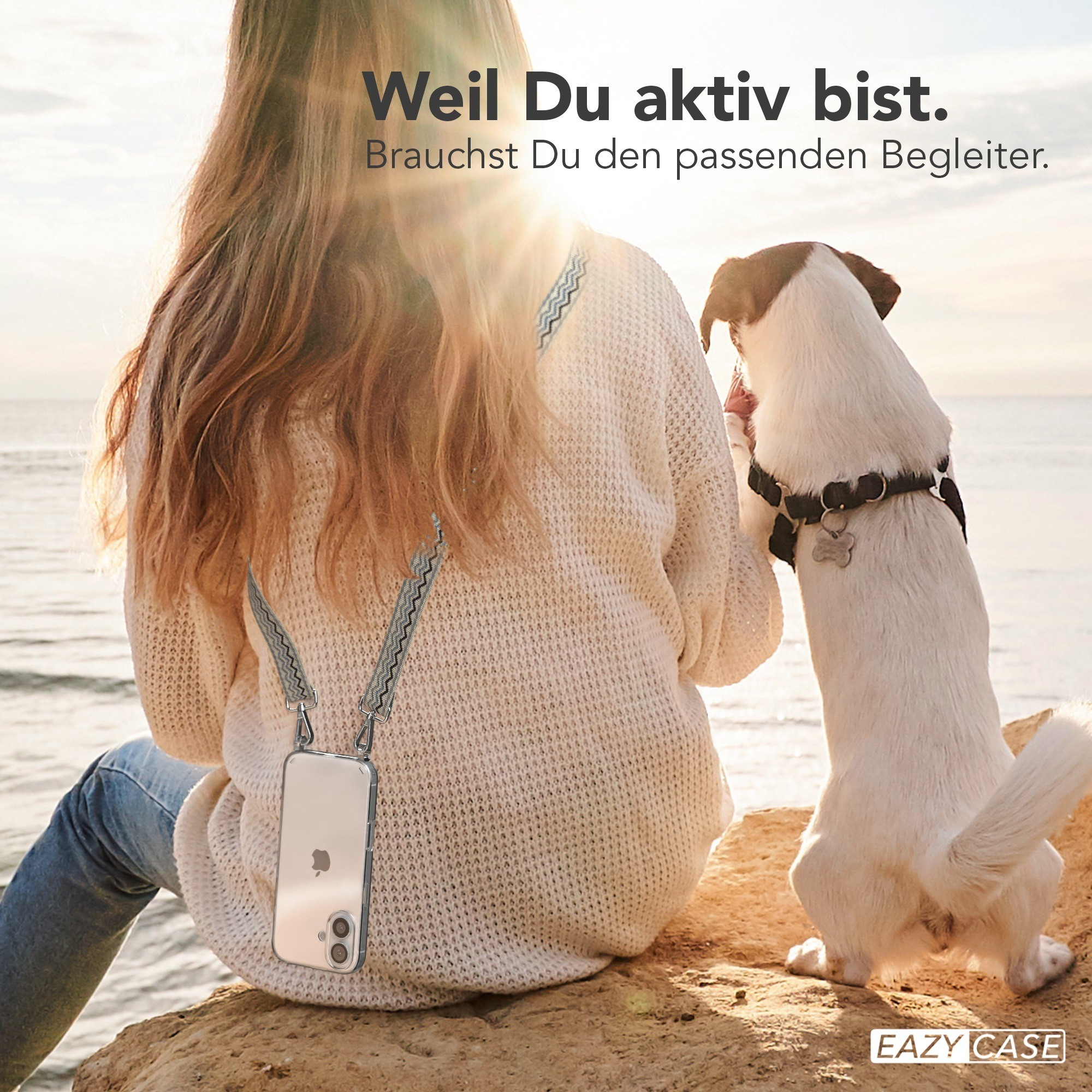 Frau mit Handyhülle und Hund am Strand. Sonne scheint. Handyhülle hat einen Trageriemen.