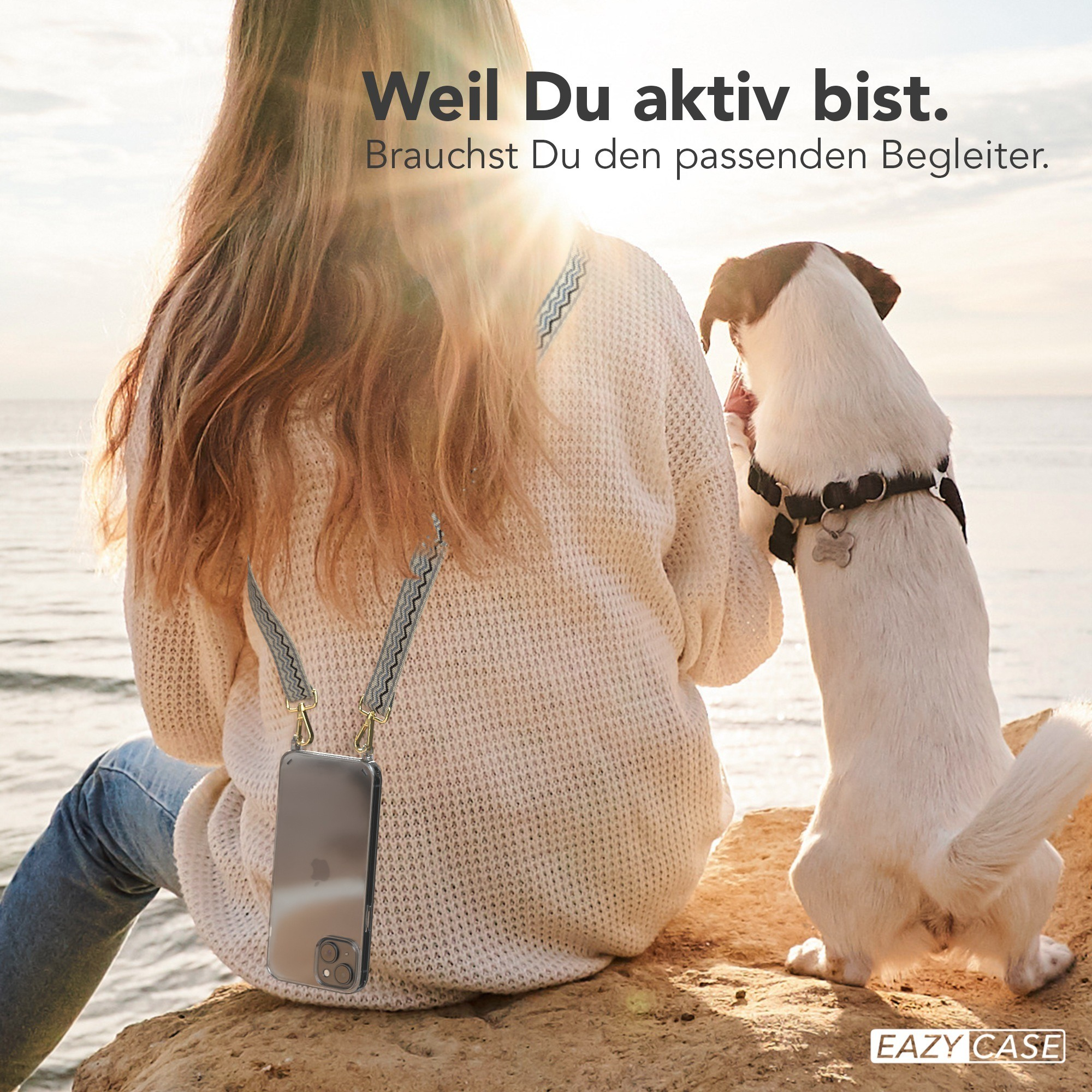 Frau sitzt mit Hund auf Felsen am Meer, hält Telefon.