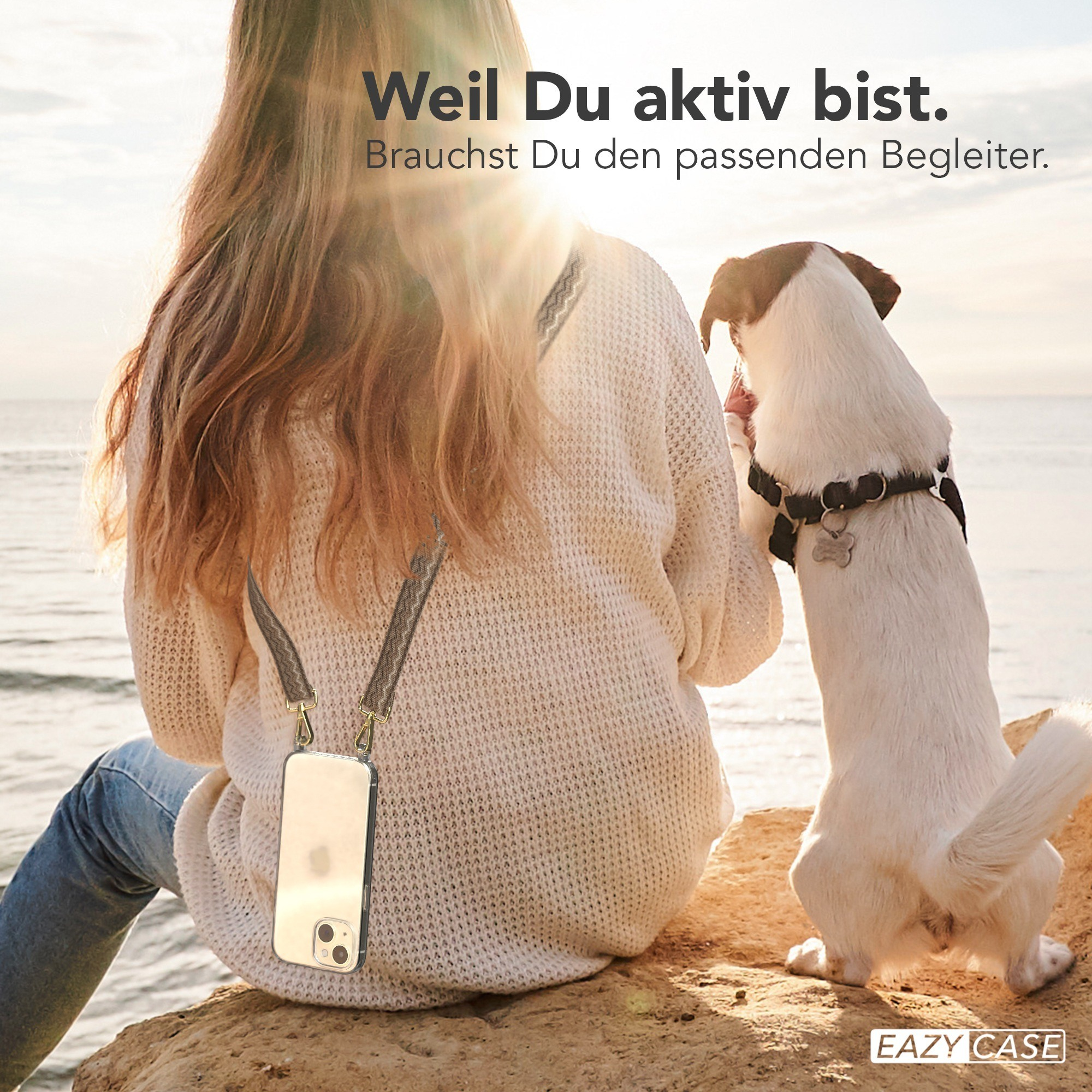 Eine Frau und ein Hund sind am Strand. Die Frau trägt ein Telefon mit einem Riemen.