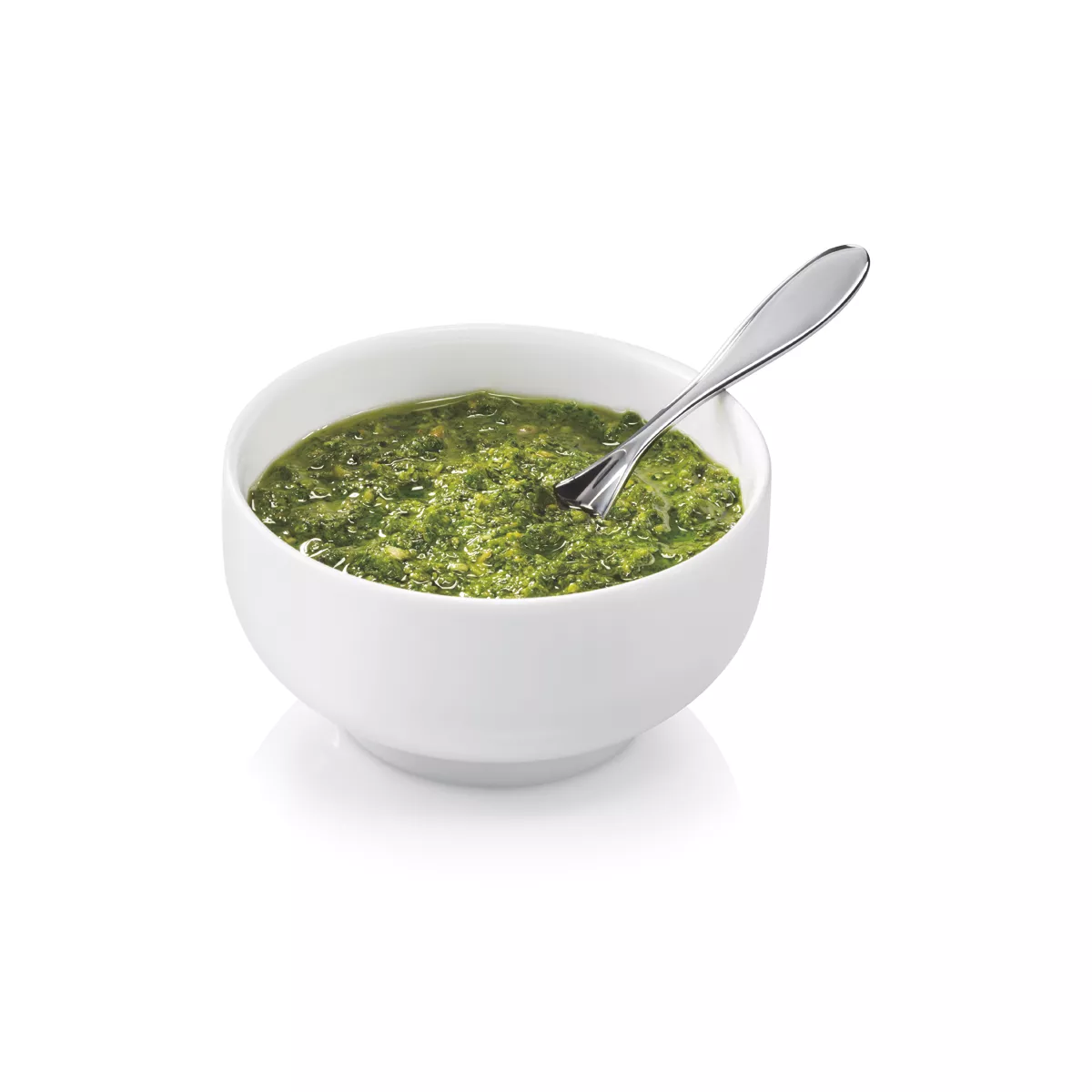 Un cuenco blanco con pesto verde y una cuchara plateada sobre una superficie blanca.