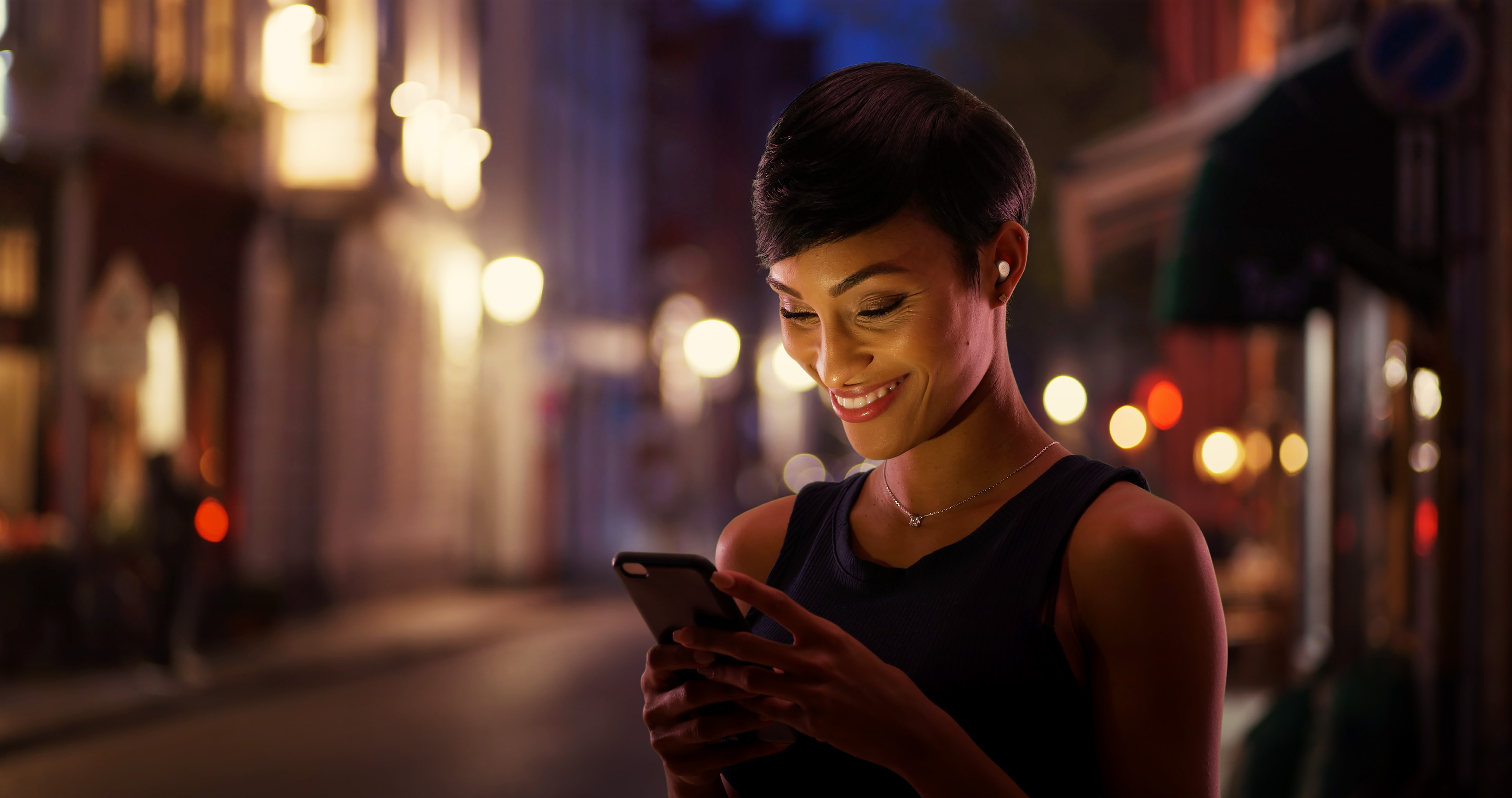Donna che usa il telefono, sorridente, con auricolari, in strada. Lo sfondo ha luci ed edifici.