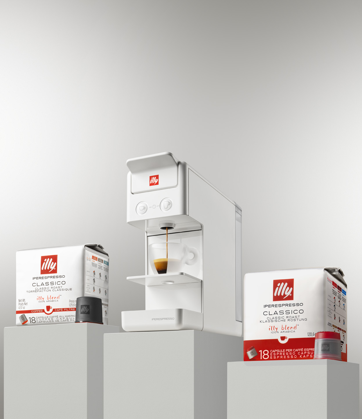 Macchina per caffè espresso bianca, con caffè versato in un bicchiere. Capsule e scatole Illy. Sfondo bianco.
