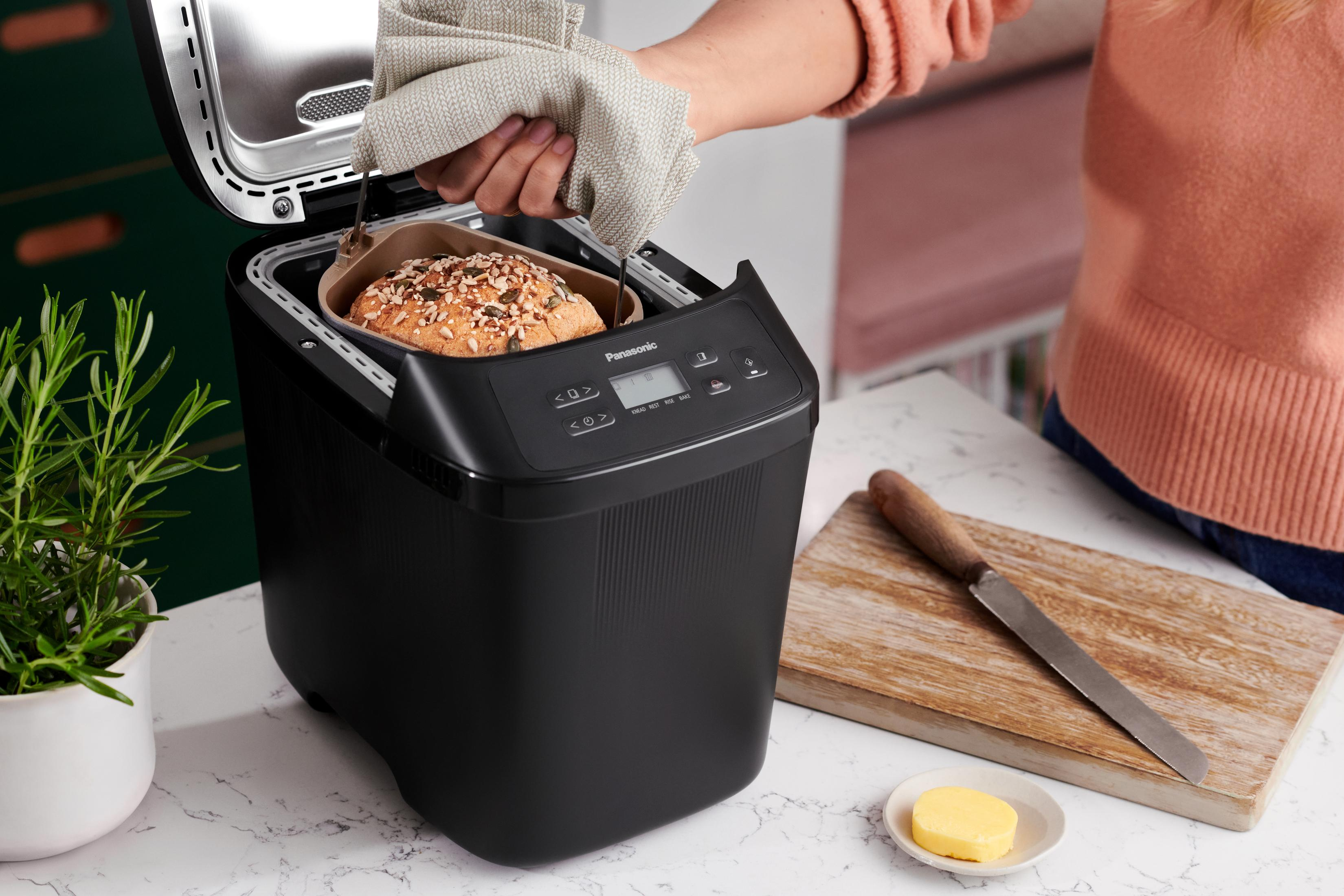 Eine Person nimmt einen Brotlaib aus einem schwarzen Brotbackautomaten. Butter und Messer daneben.