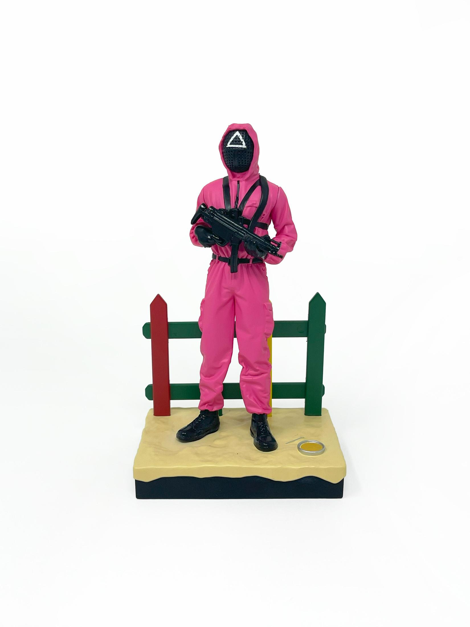 Figura de El Juego del Calamar. Mono rosa, máscara negra, sosteniendo una pistola. Se para sobre arena.