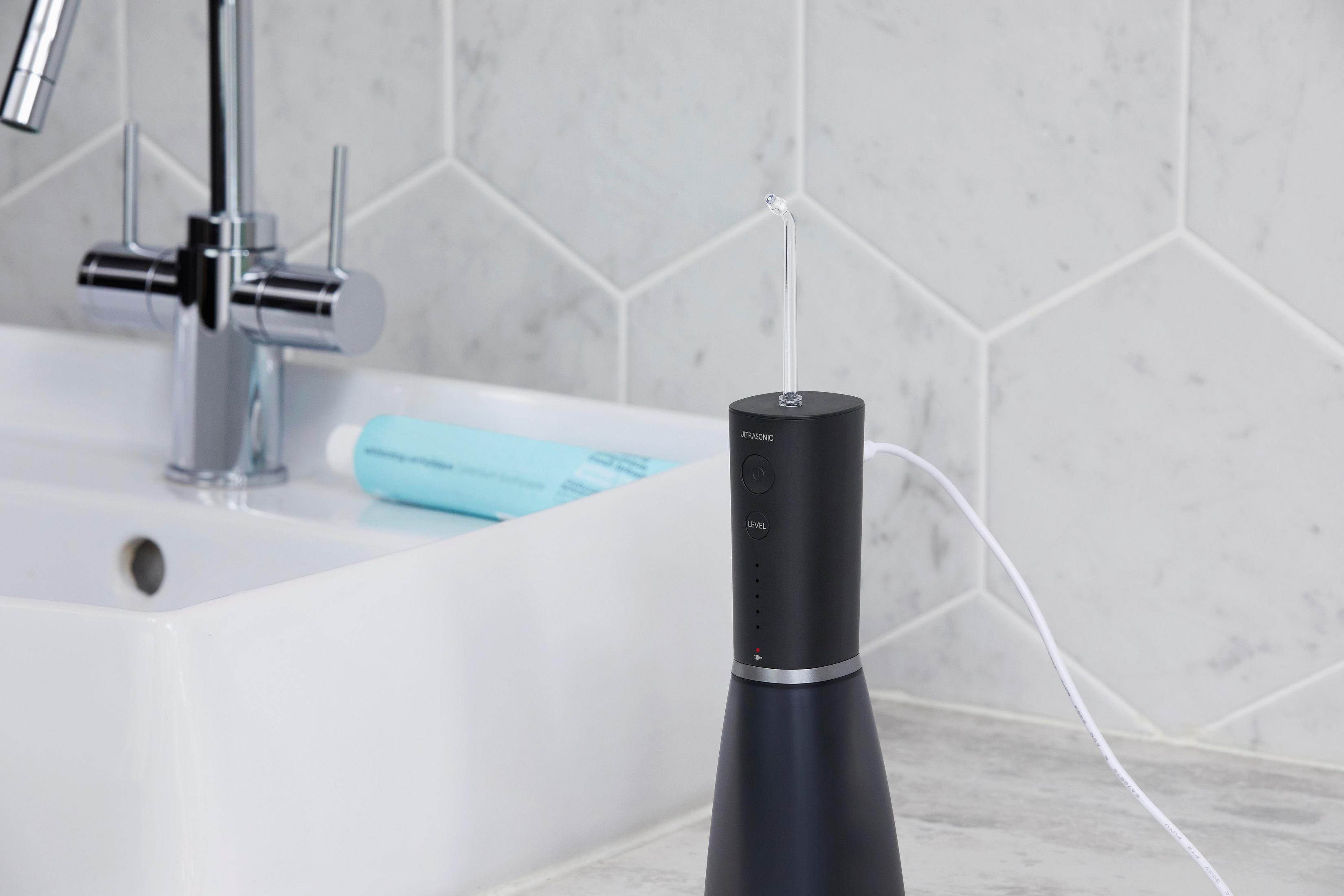 Black and silver water flosser on a white sink, tap in the background. White tiles on the wall.