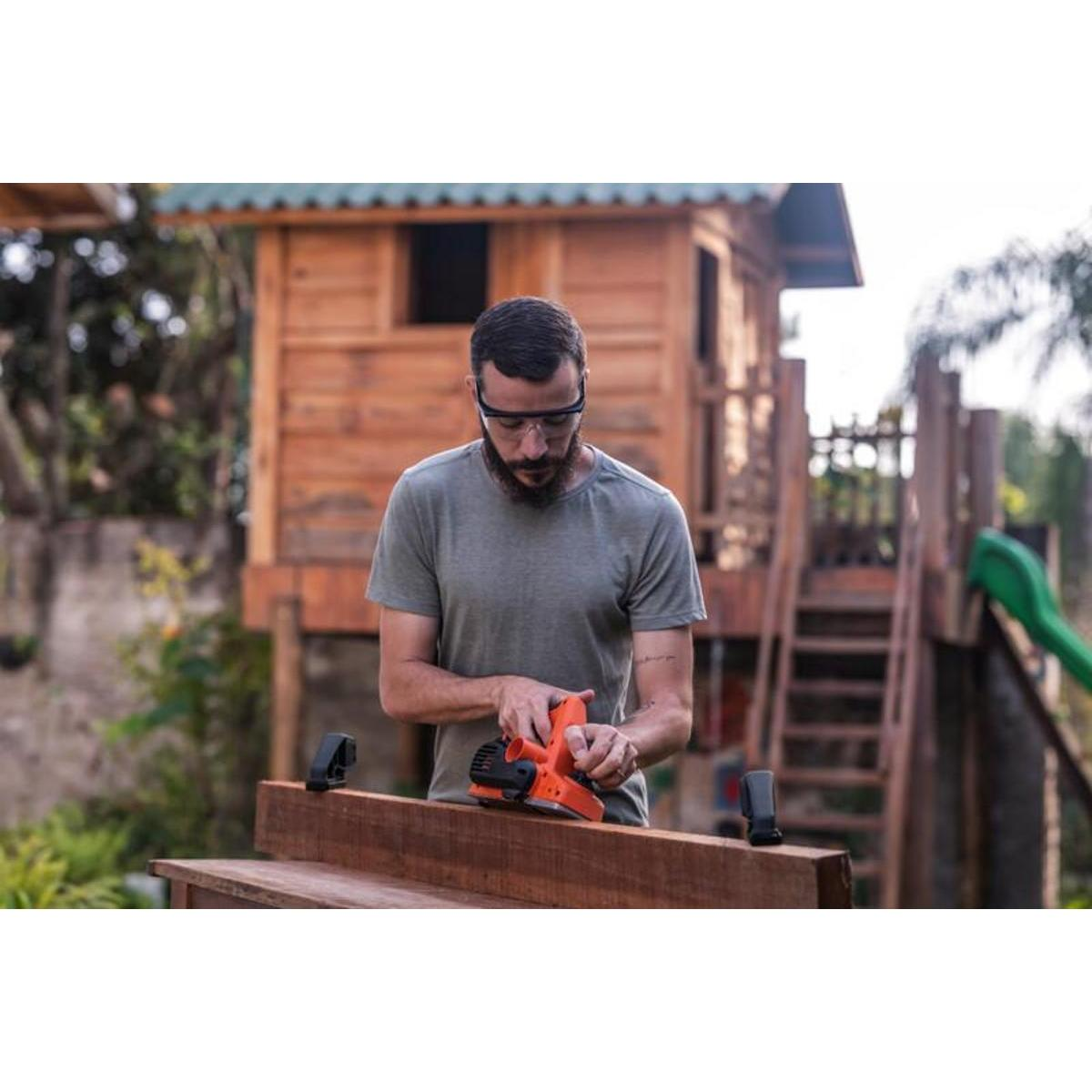 Un hombre con gafas cepilla una tabla de madera con una cepilladora naranja. Una casita de madera al fondo.