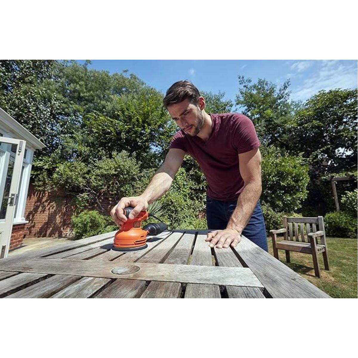 Mann schleift im Freien einen Holztisch mit einem orange-schwarzen Schleifer.