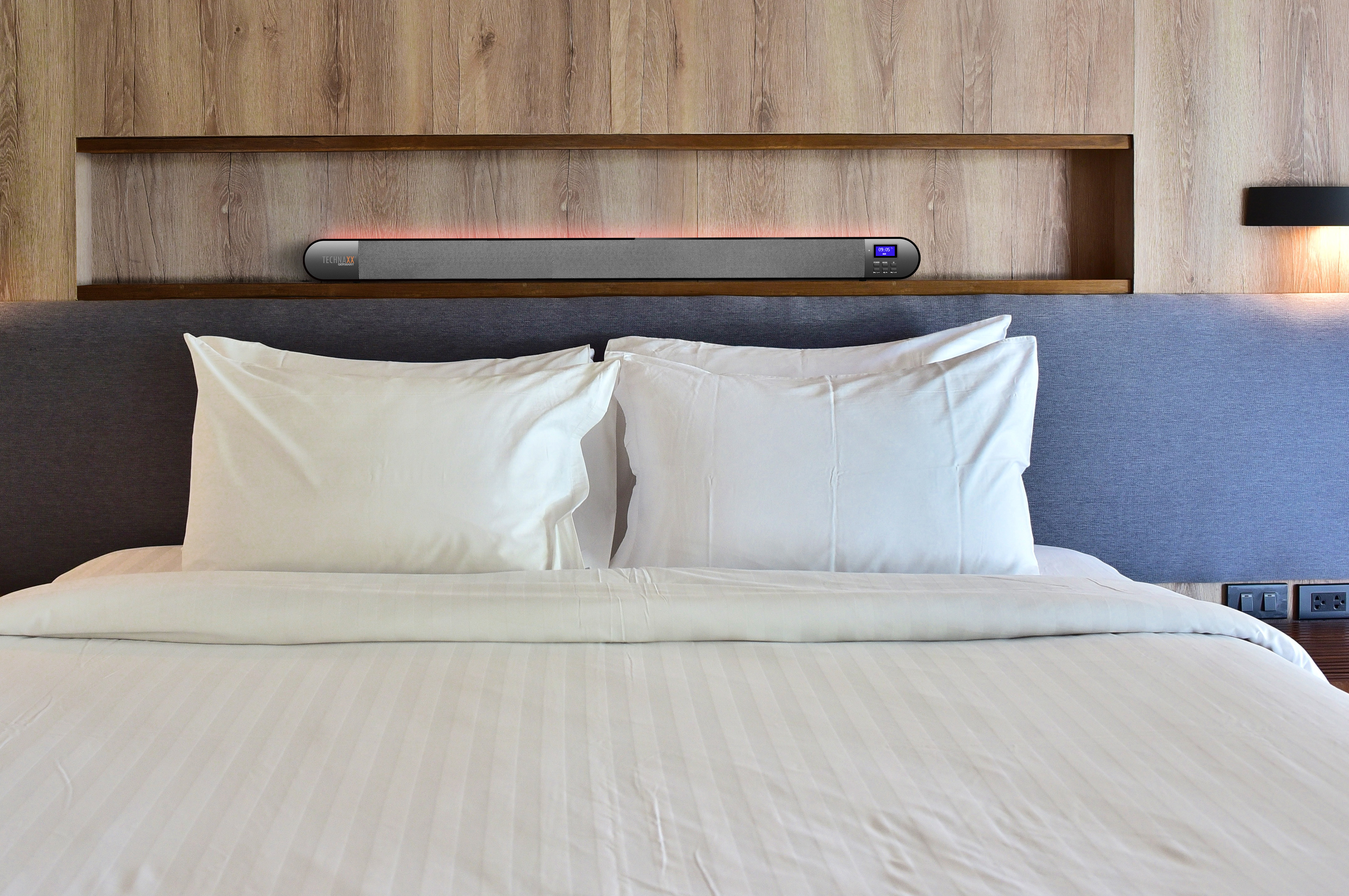 Una soundbar rettangolare è posizionata su una mensola sopra un letto. Due cuscini sono sotto.