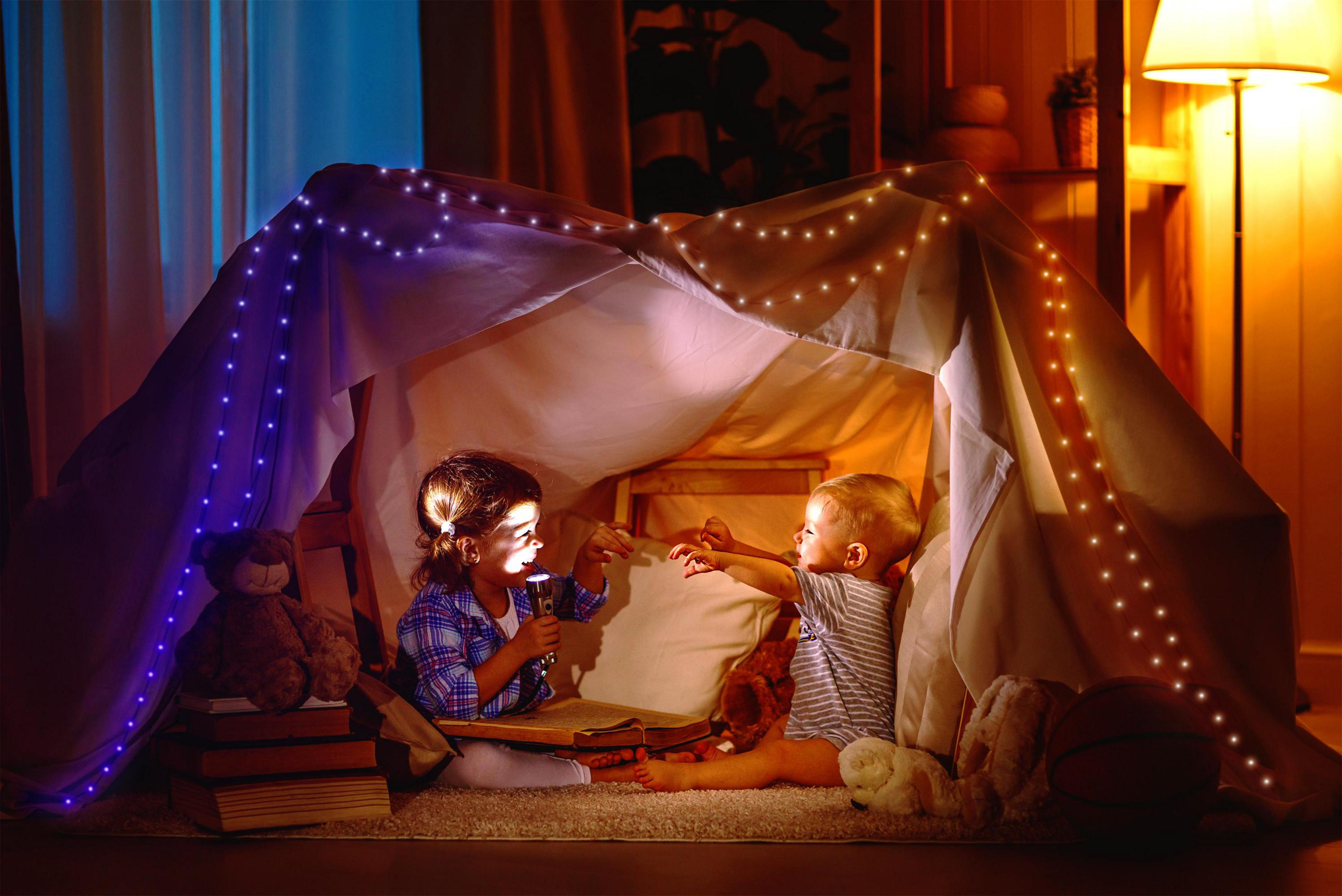 Dwoje dzieci w namiocie, jedno z latarką, czytają książkę.