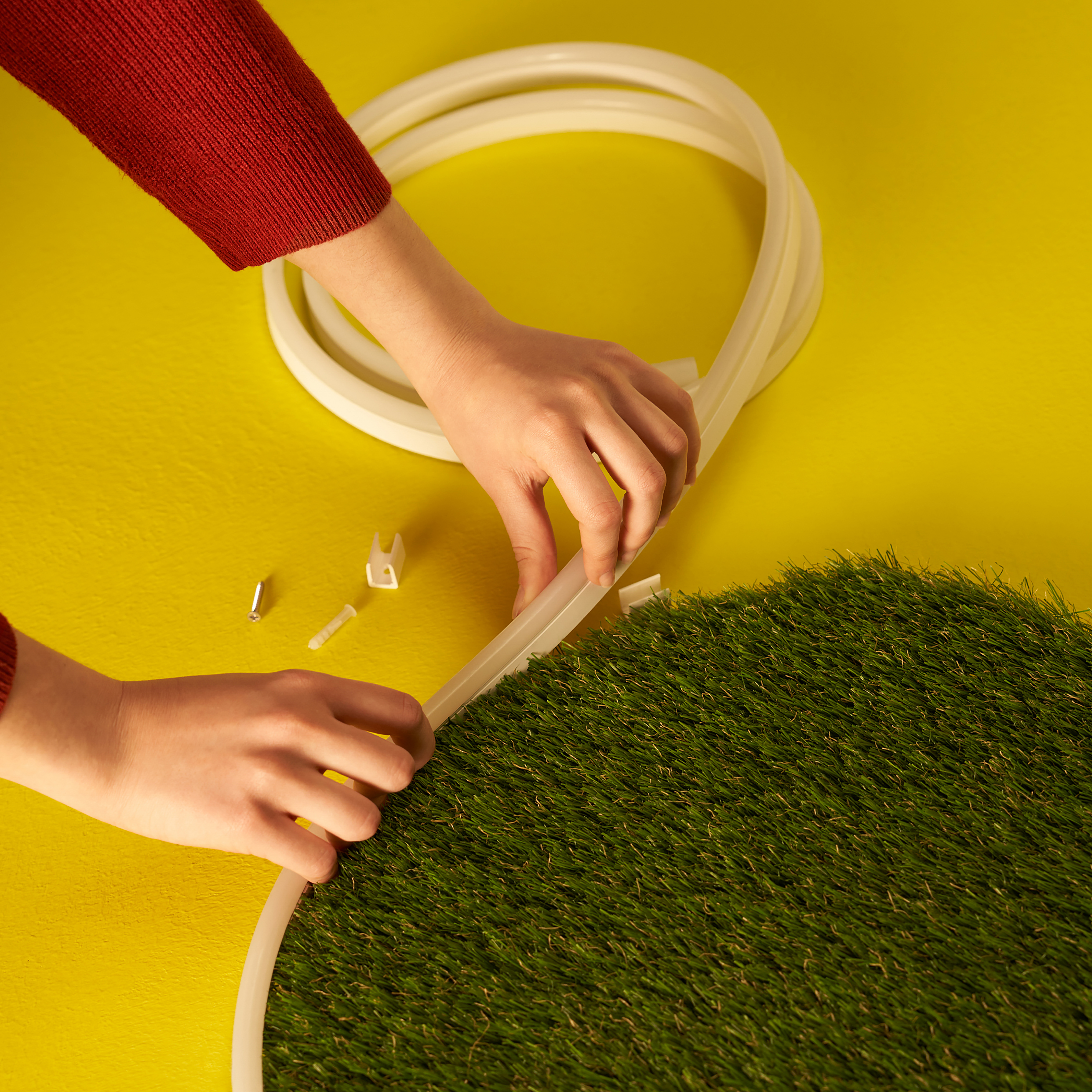Hands assembling a circular strip of artificial turf. A yellow surface forms the background.