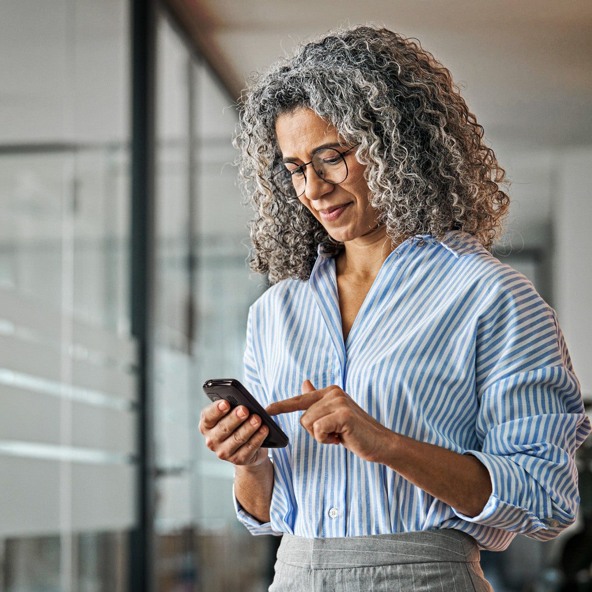 Donna con occhiali sorride, usa un telefono. Indossa una camicia a righe e una gonna grigia. Sfondo con pannelli di vetro.