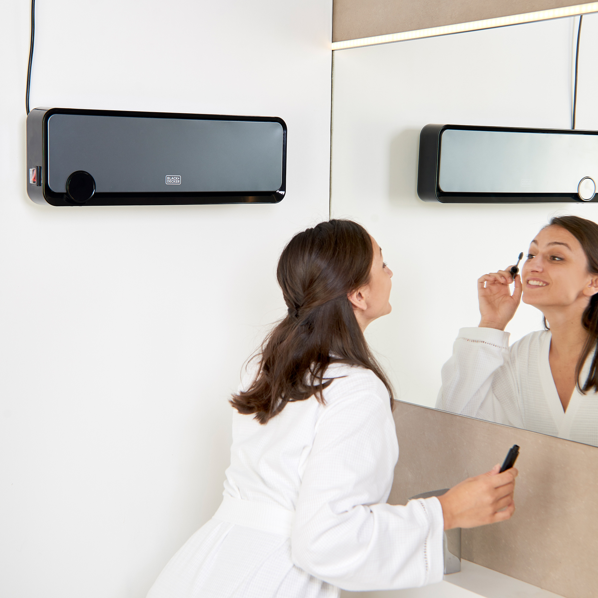 Kobieta w szlafroku nakłada makijaż przed lustrem. Czarny grzejnik na ścianie.