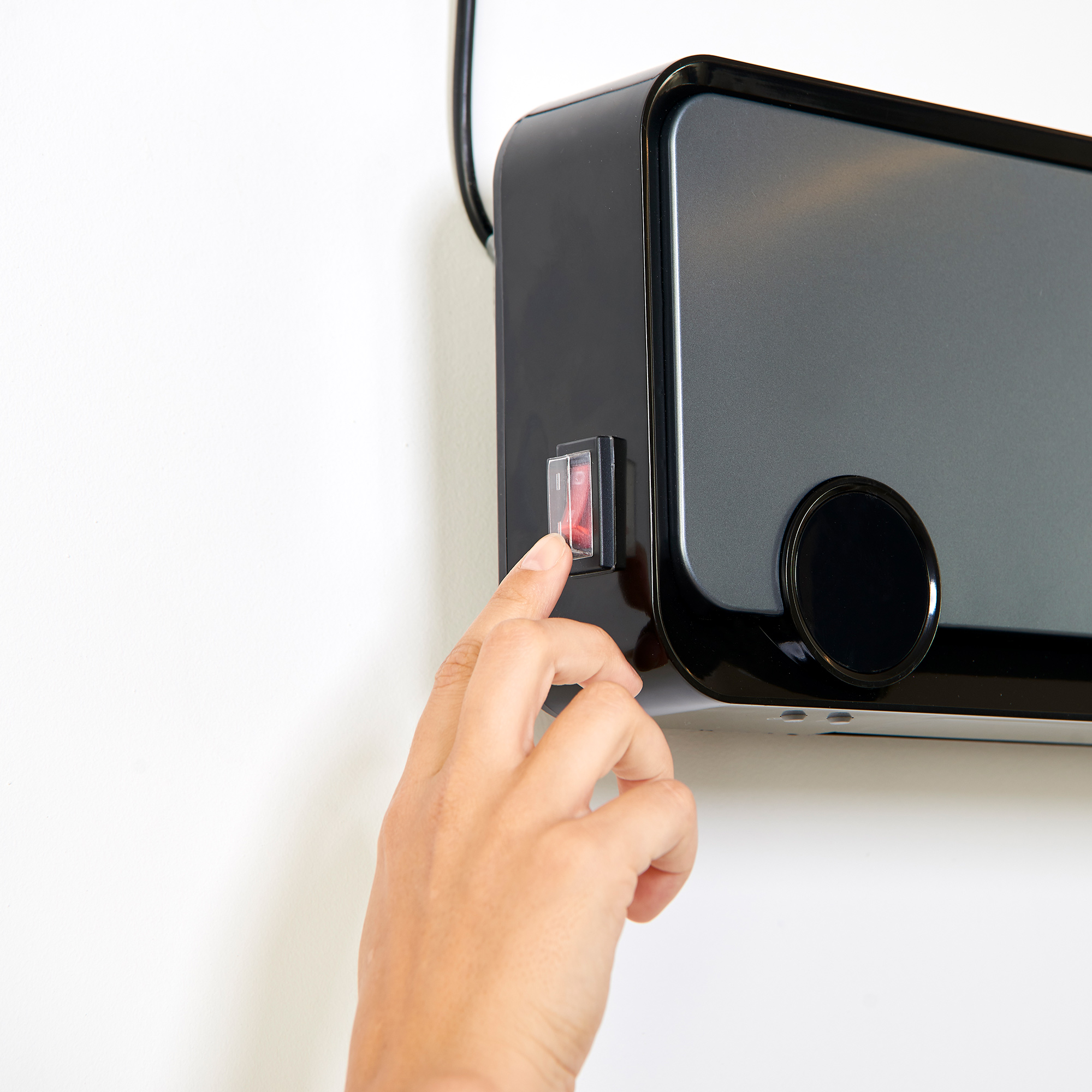 Dłoń włącza czarny, montowany na ścianie grzejnik z czerwonym przełącznikiem.