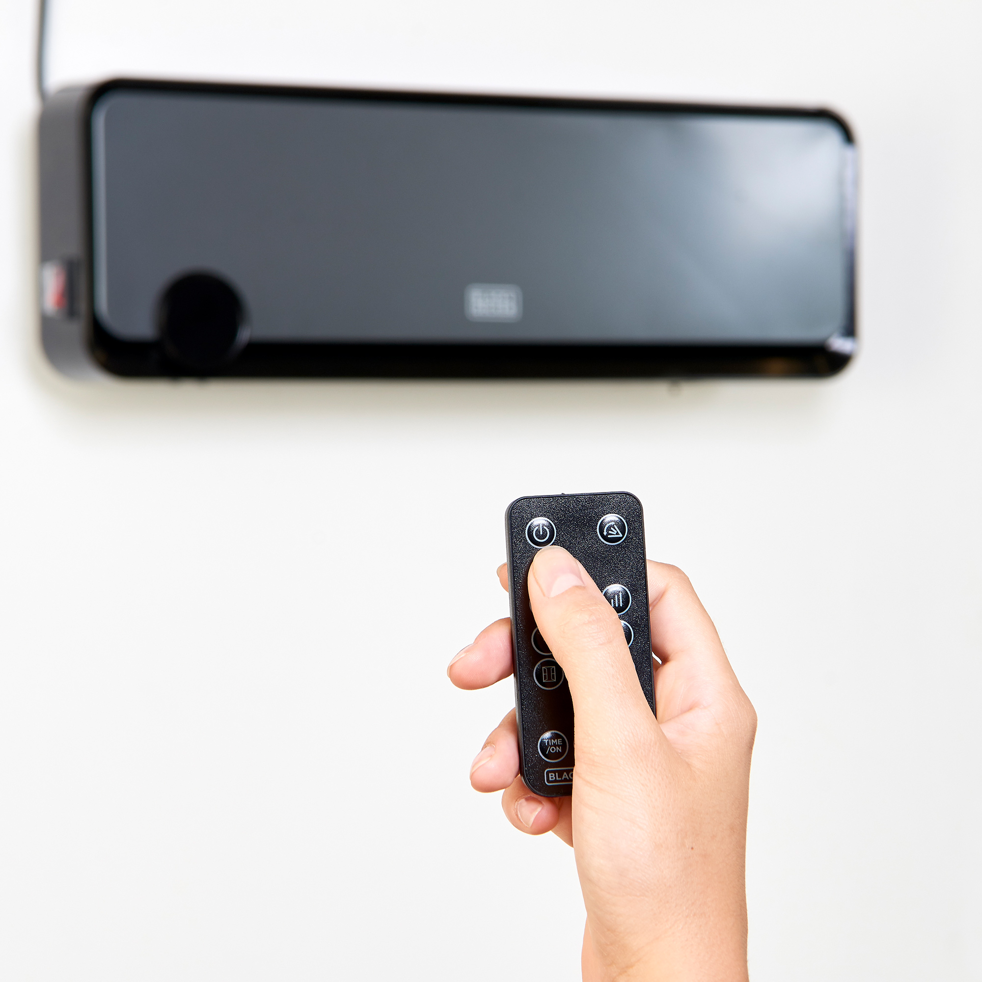 Ręka trzymająca pilota skierowanego na czarne urządzenie na białej ścianie. Logo DECKER.