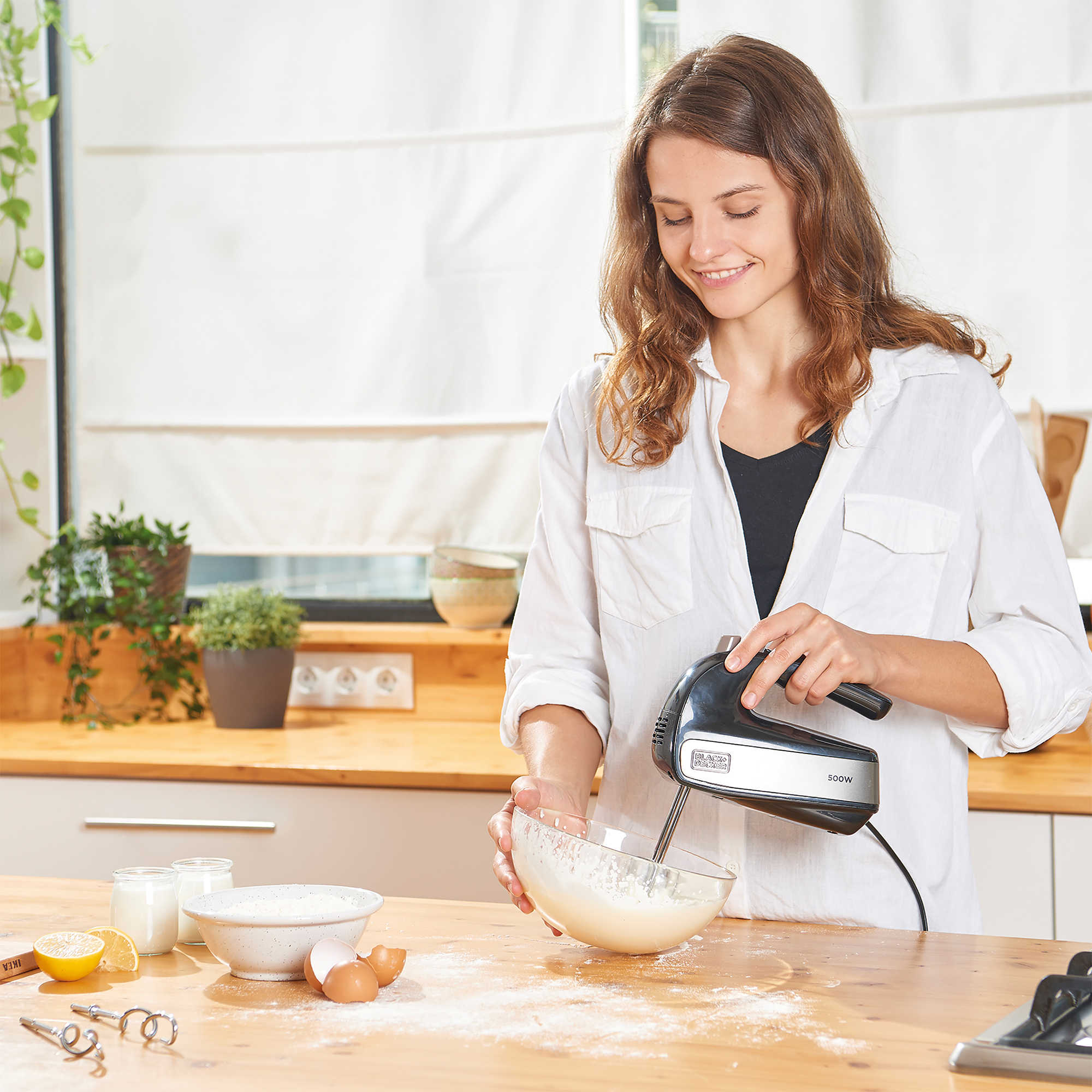 Eine Frau mischt Zutaten in einer Glasschüssel mit einem Handmixer, Küchenumgebung.
