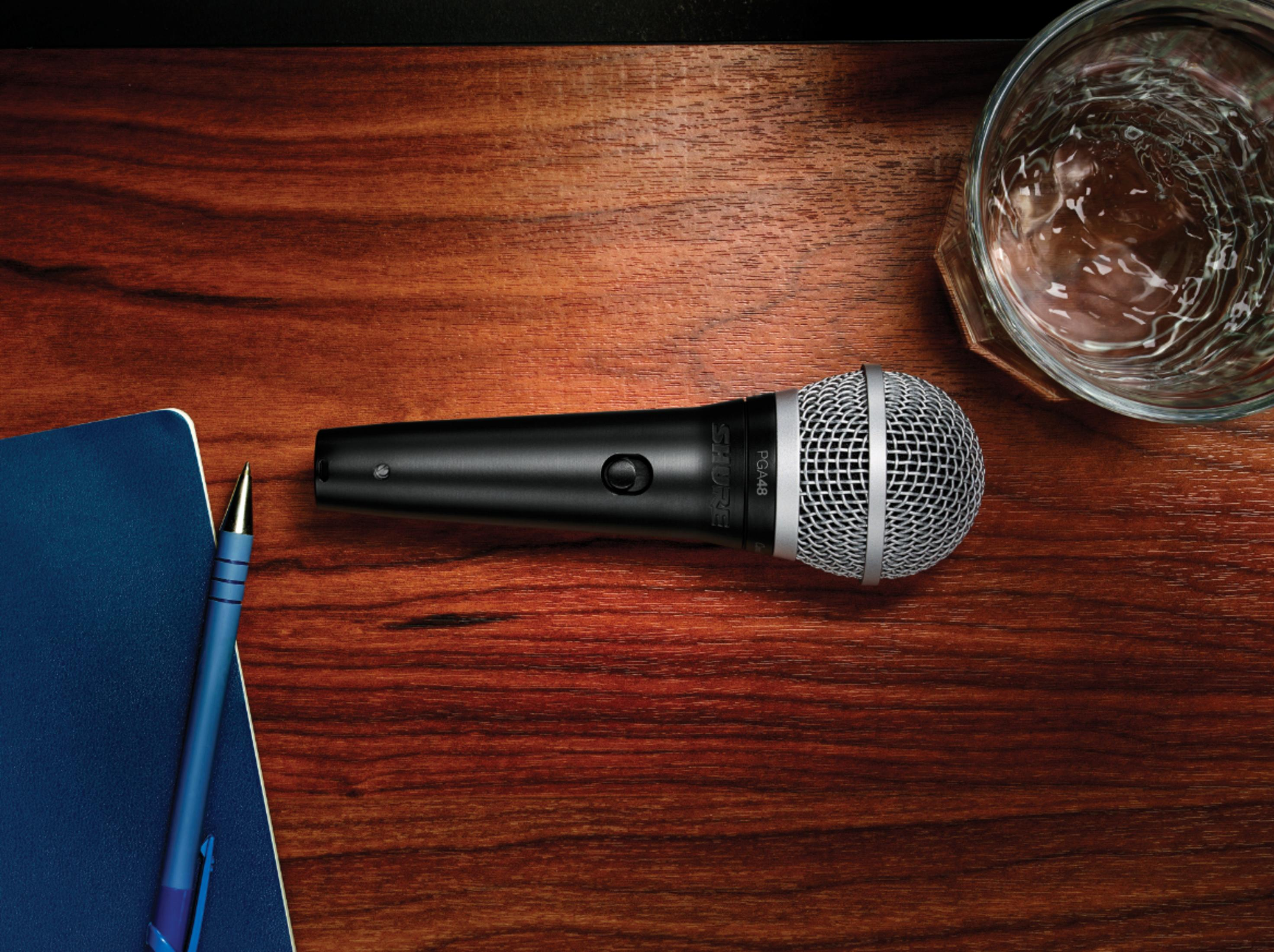 Mikrofon na drewnianym stole ze szklanką wody, długopisem i notatnikiem. Scena jest dobrze oświetlona.