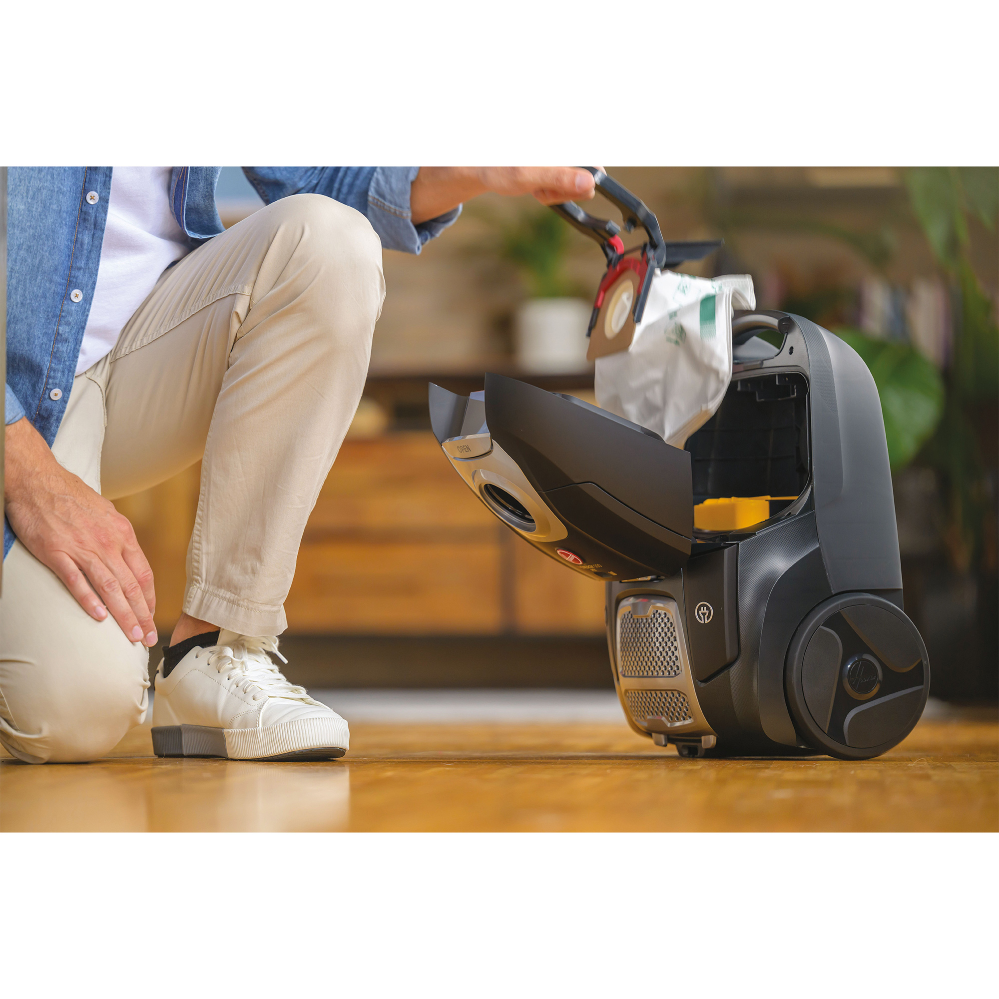 Osoba wkłada nowy worek do czarnego odkurzacza. Drewniana podłoga i szafka w tle.