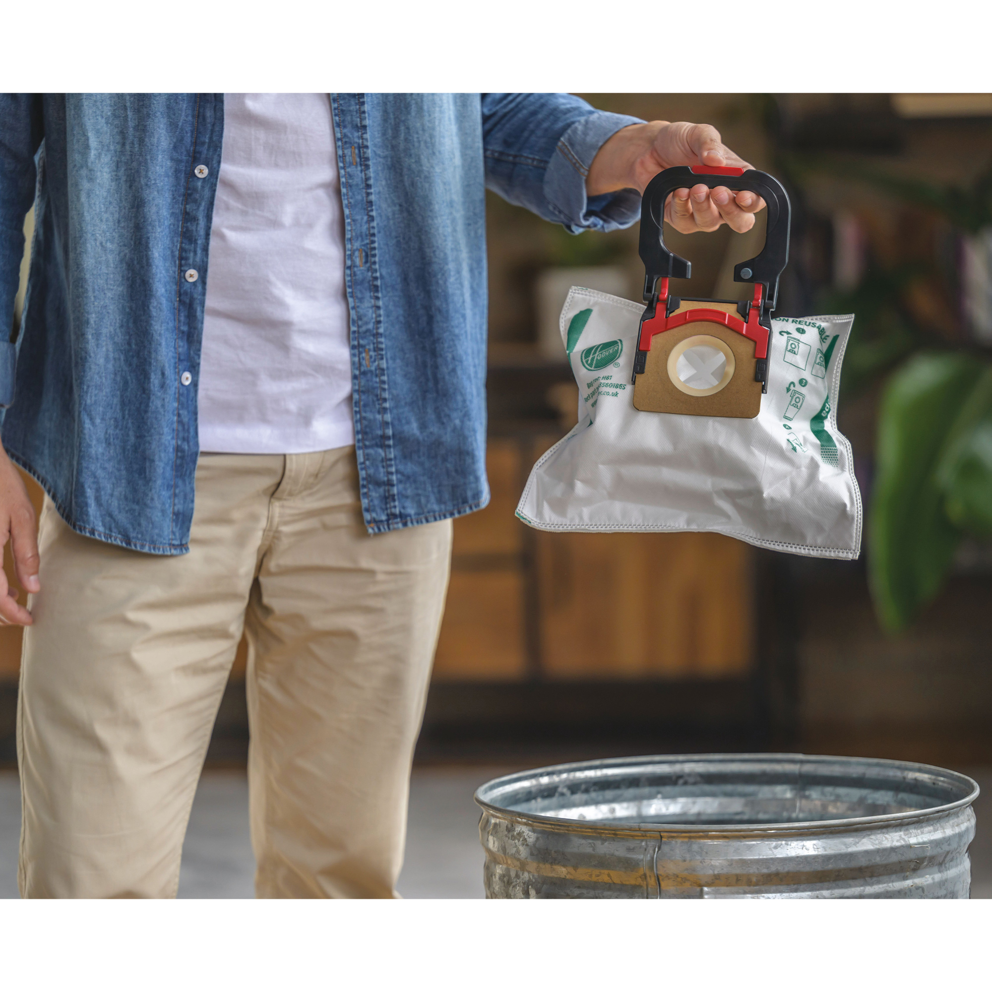 Osoba trzymająca worek do odkurzacza nad koszem na śmieci, w dżinsach i beżowych spodniach.