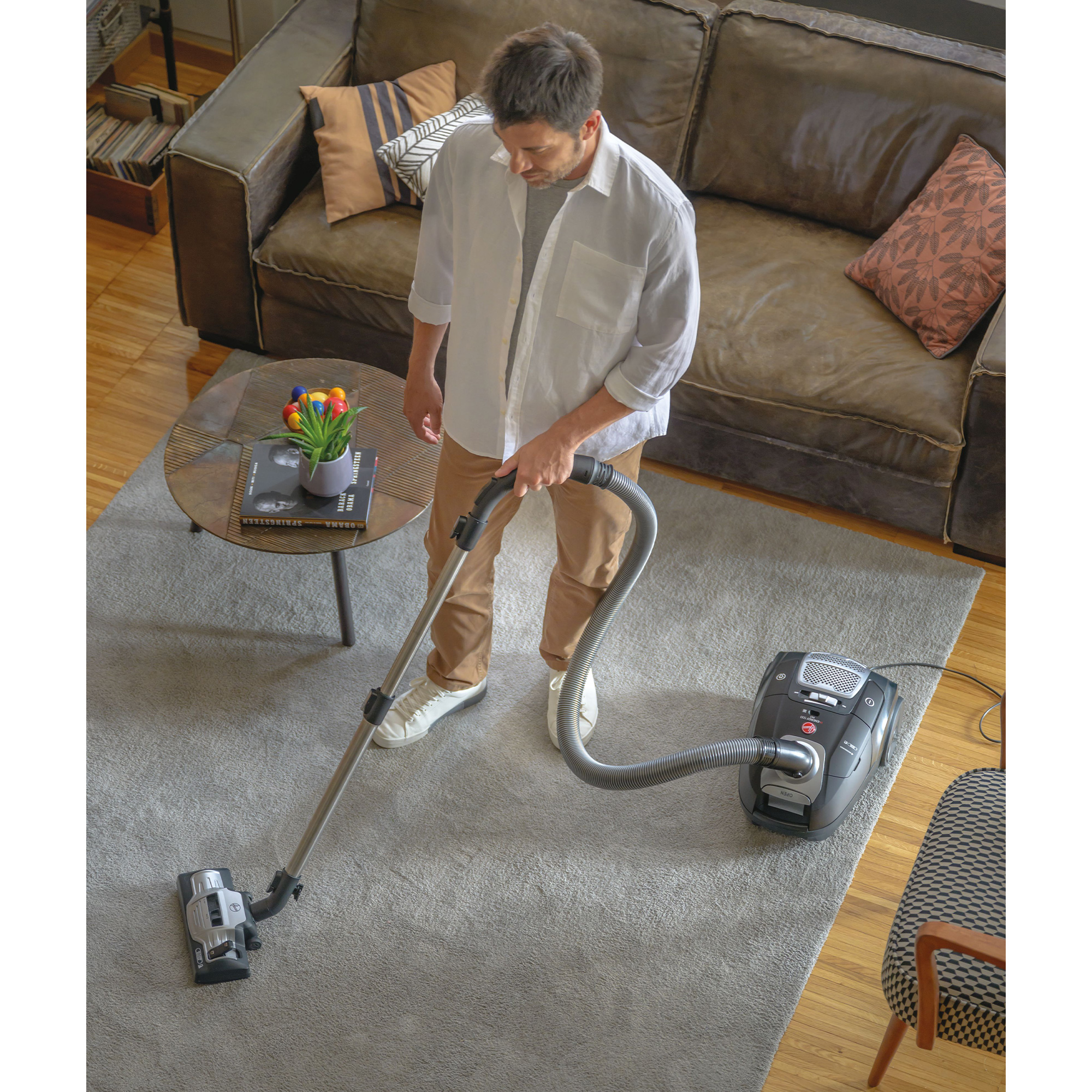 Mężczyzna odkurza dywan. W tle kanapa, stolik kawowy i dywan. Odkurzacz jest czarny.