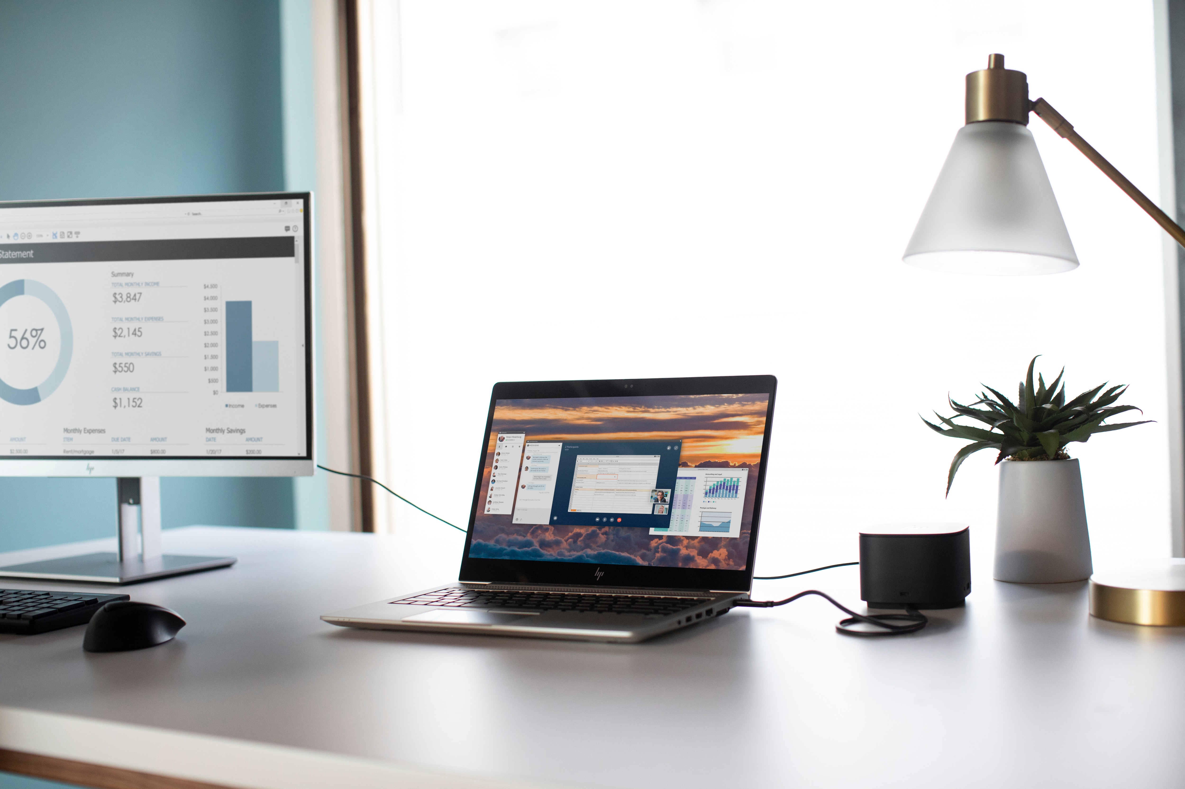 Ustawienia biurka z laptopem, monitorem, lampą i rośliną. Ekran z wykresami.