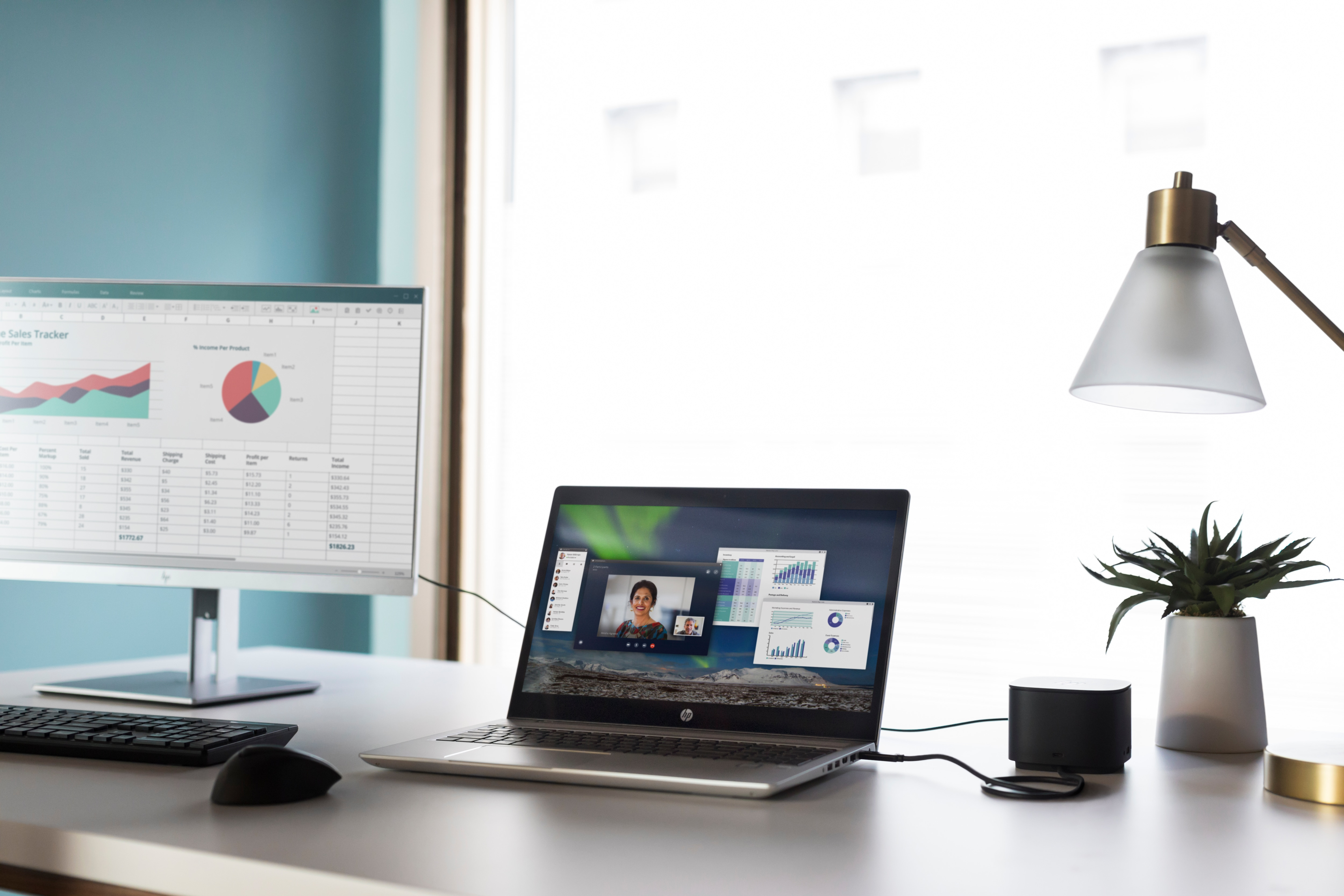 Laptop i monitor na biurku, pokazujące rozmowę wideo i arkusze kalkulacyjne, blisko okna.
