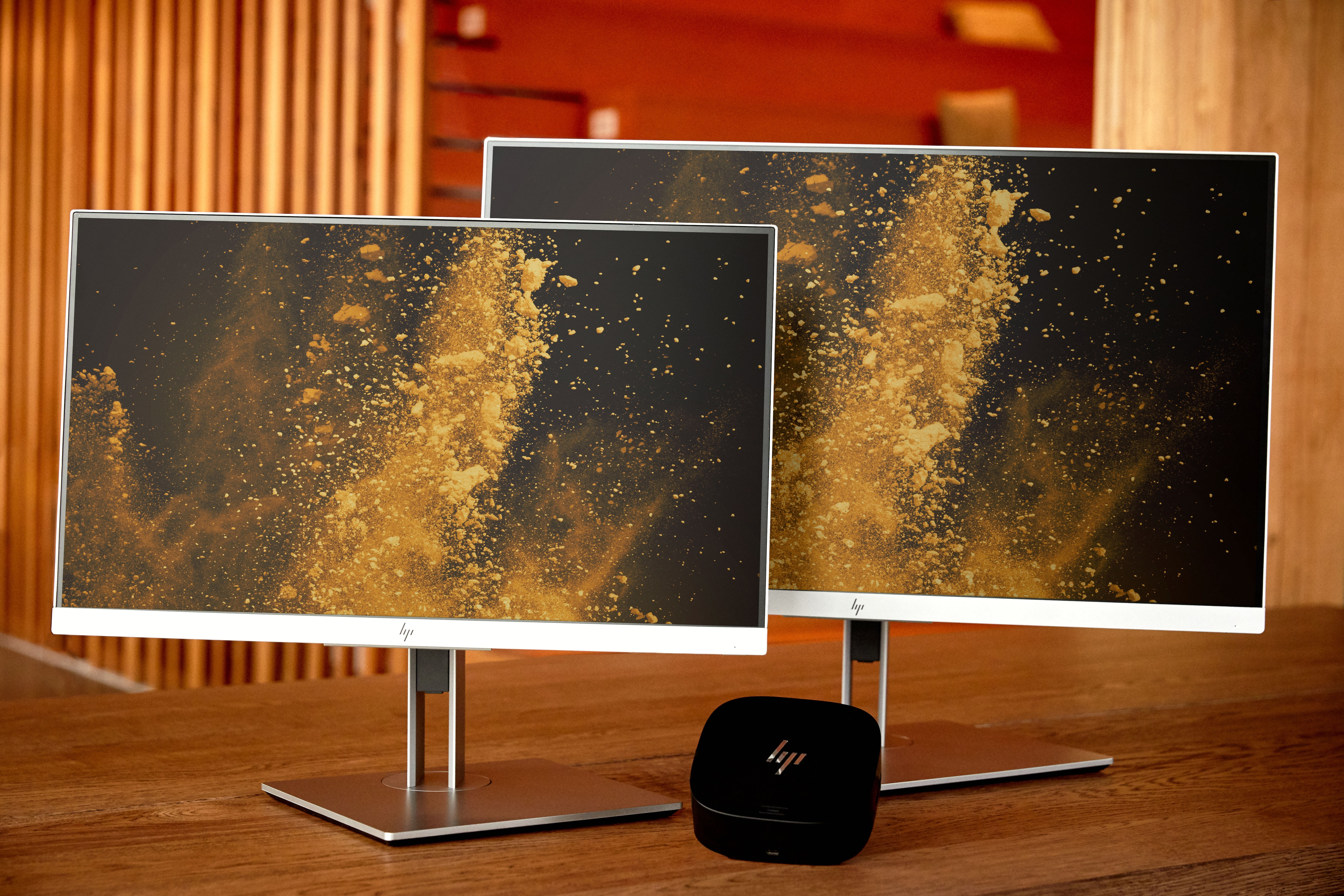 Due monitor e un piccolo dispositivo nero su un tavolo di legno, con sfondo arancione.