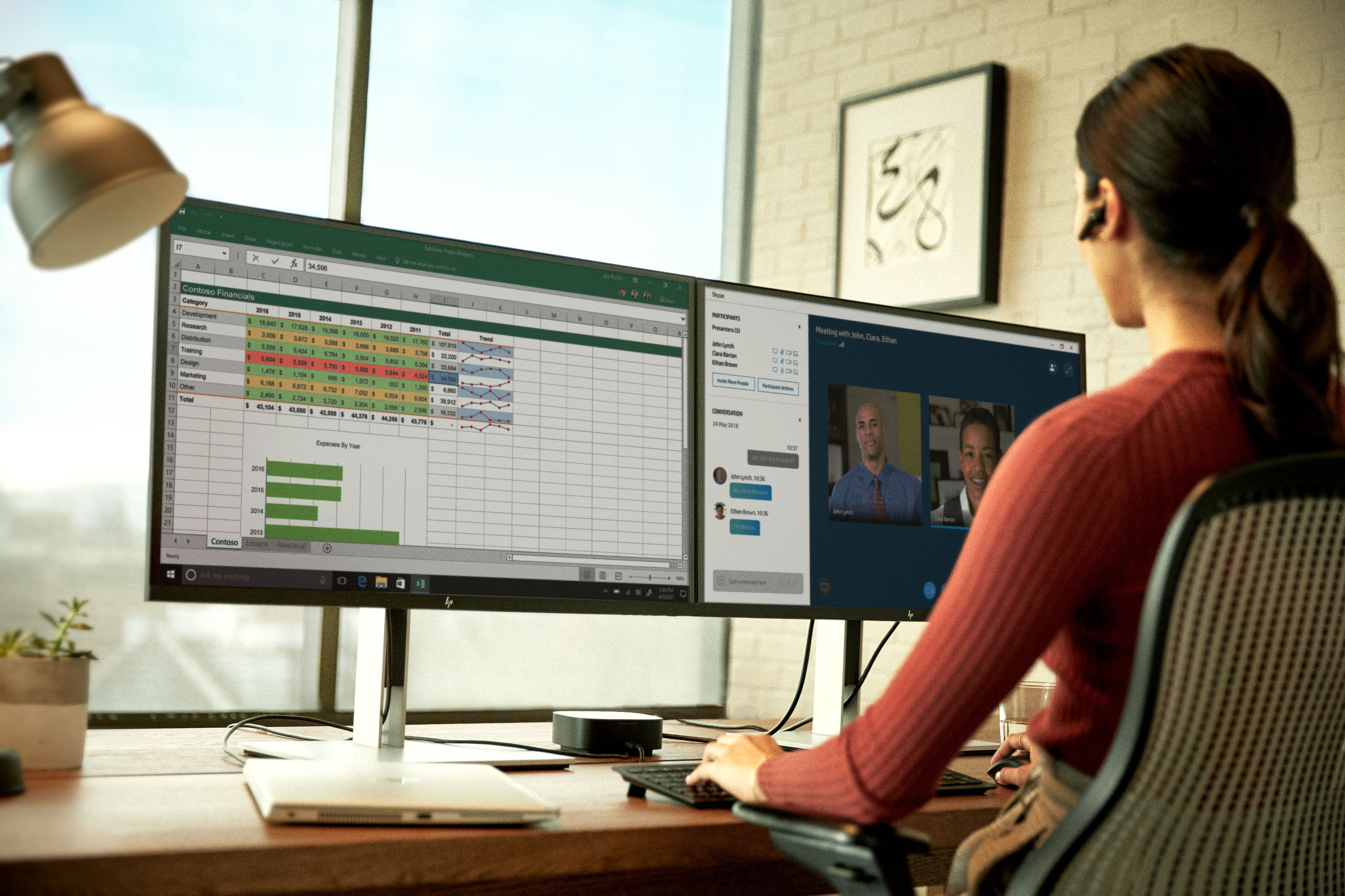 Donna che utilizza doppi monitor con Excel e videochiamata. Laptop, lampada da scrivania e sedia. Parete in mattoni.
