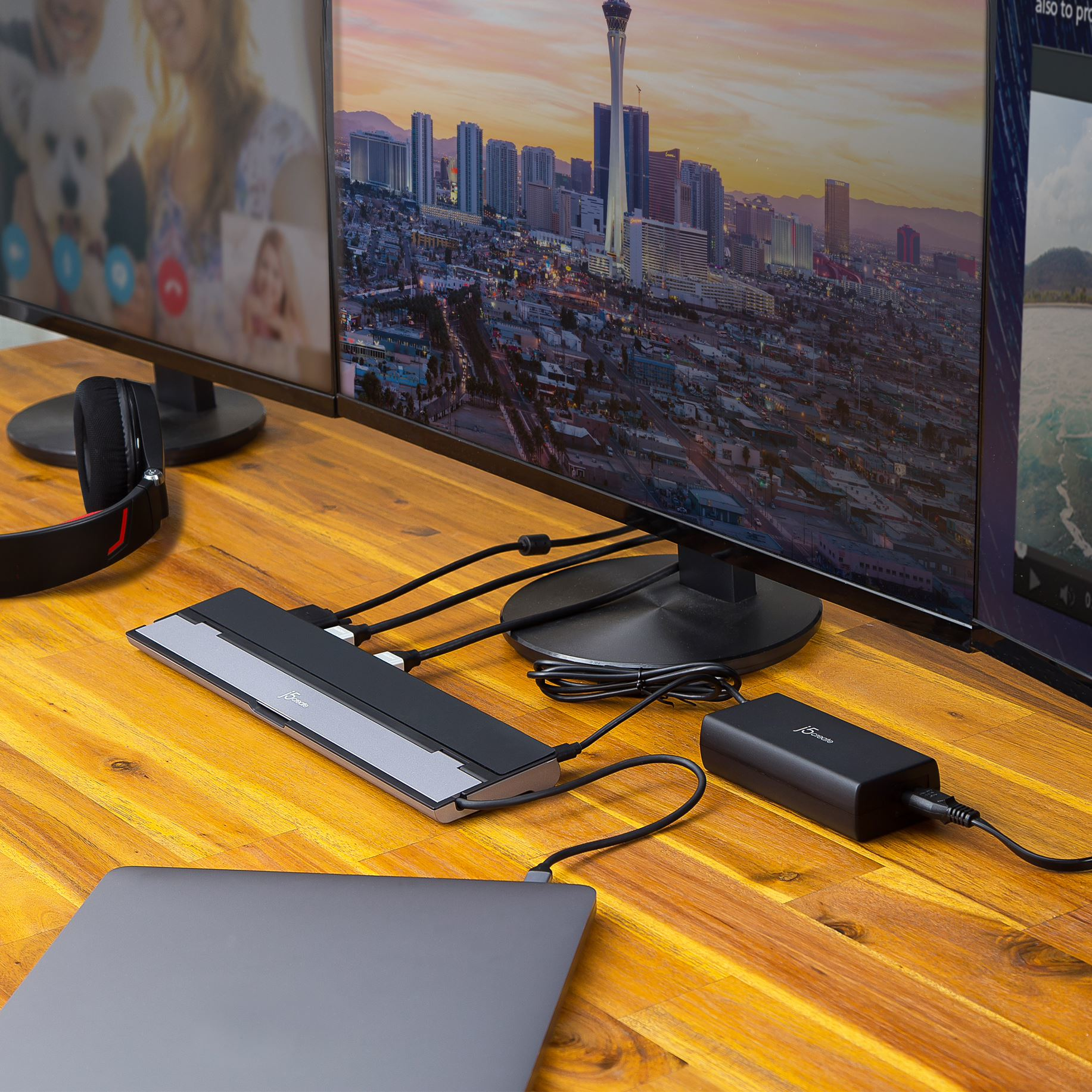 Un laptop e due monitor su un tavolo, collegati a un hub e un adattatore di alimentazione. Lo sfondo mostra un paesaggio urbano.