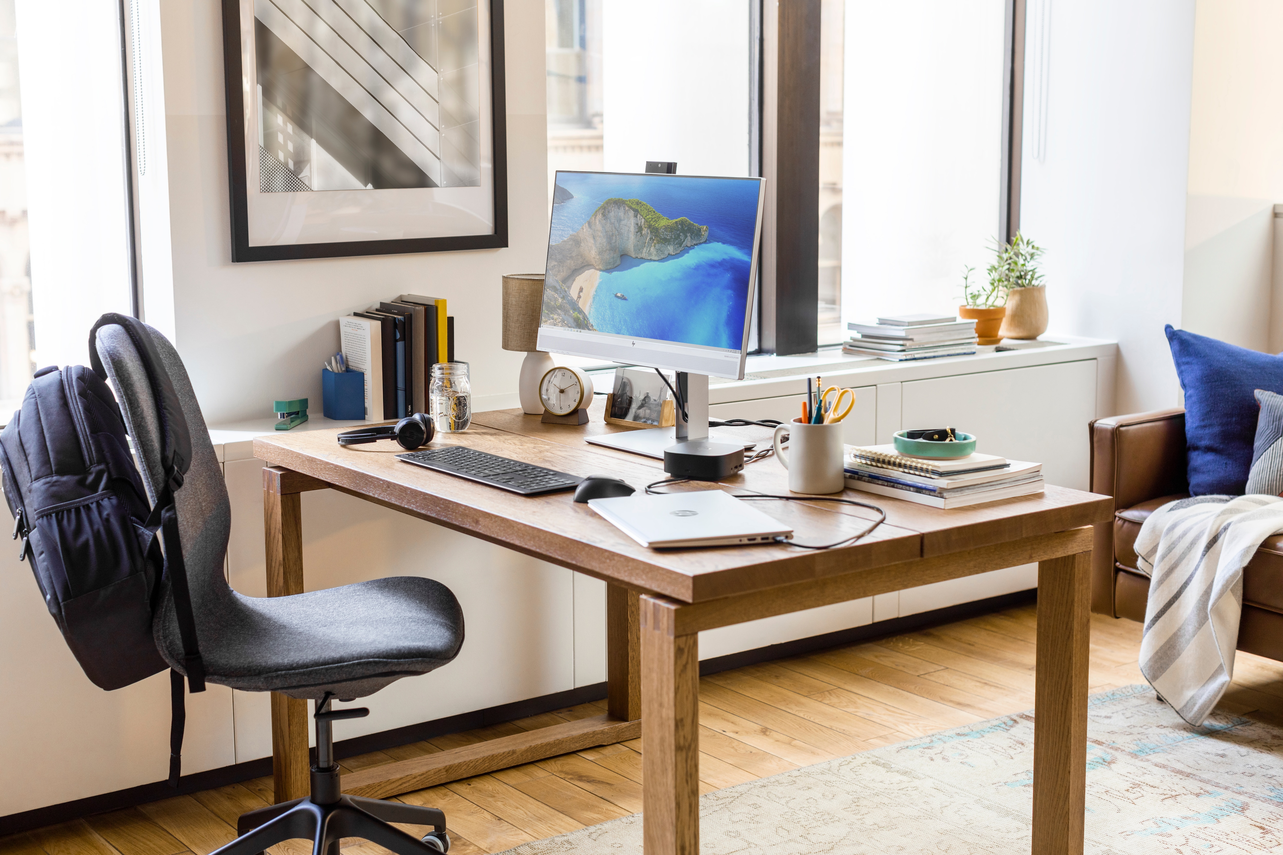 Schreibtisch mit Computer, Büchern und Rucksack auf einem Stuhl. Fenster mit Tageslicht.