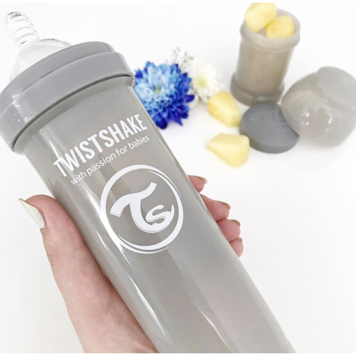 Eine graue Babyflasche mit Twistshake-Etikett, gehalten von einer Hand, mit Blumen und anderen Gegenständen im Hintergrund.