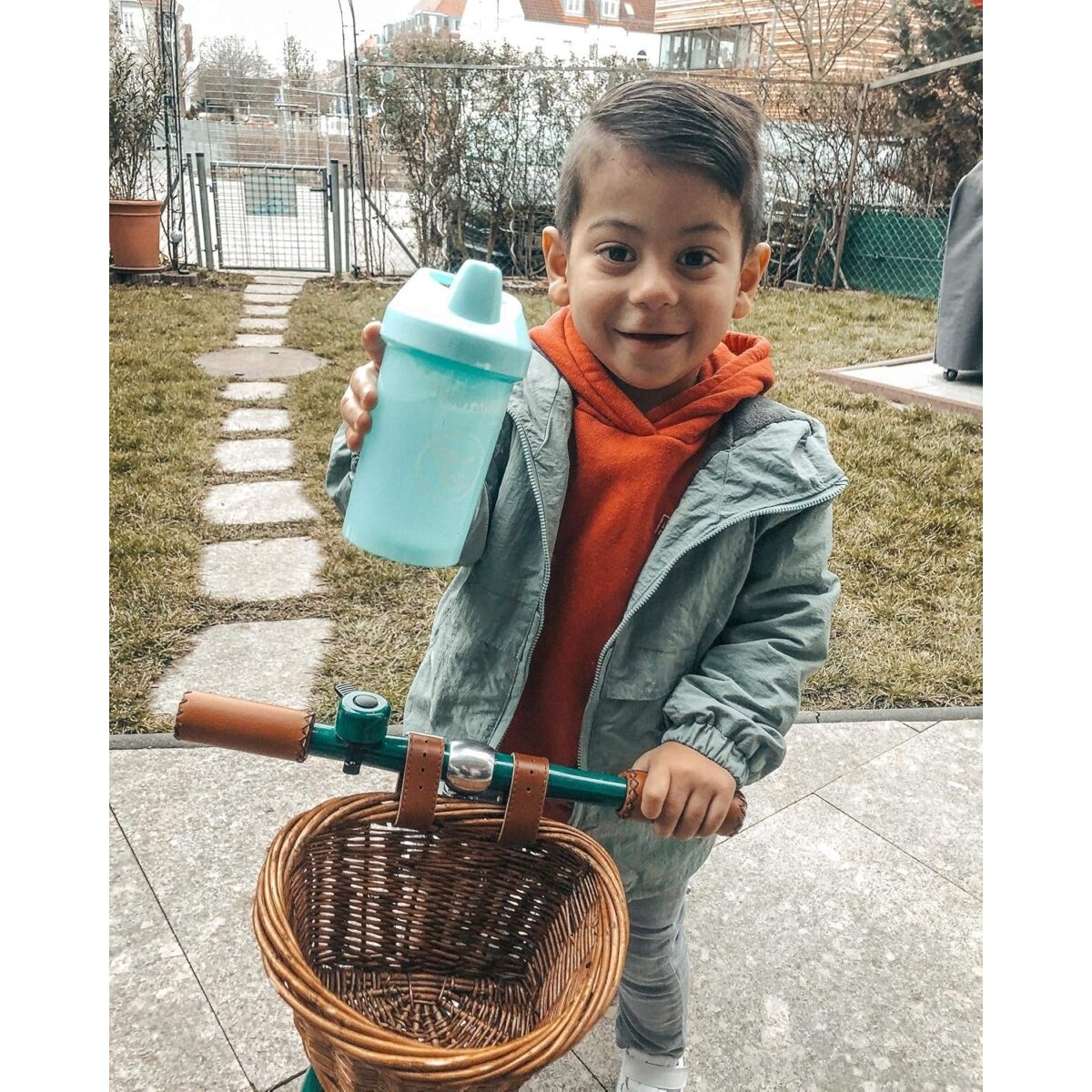 Junge hält einen blauen Trinkbecher, mit Roller, auf einem gepflasterten Weg.