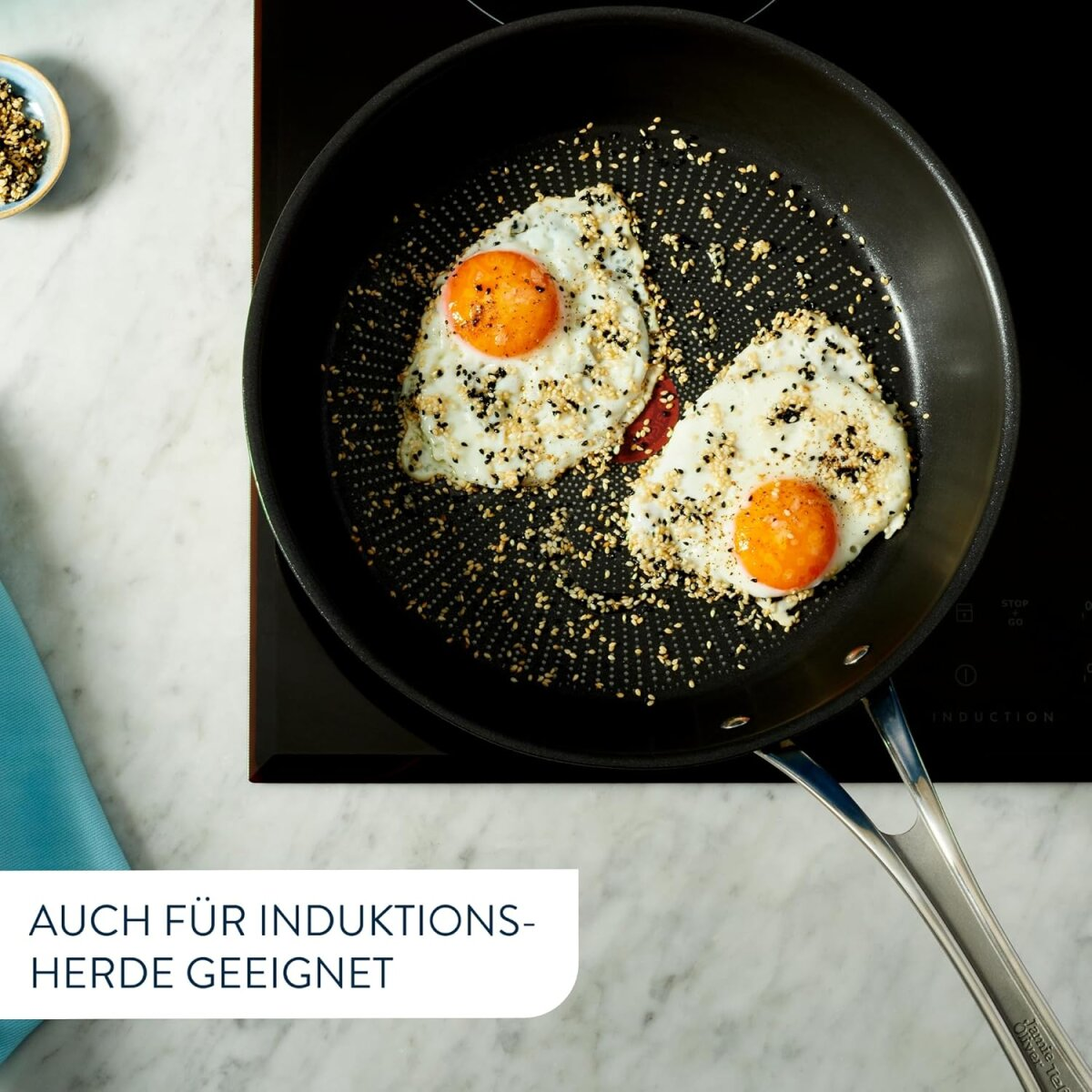 Zwei Spiegeleier in einer schwarzen Pfanne, mit Sesam. Text sagt: 'auch für Induktionsherde geeignet'.