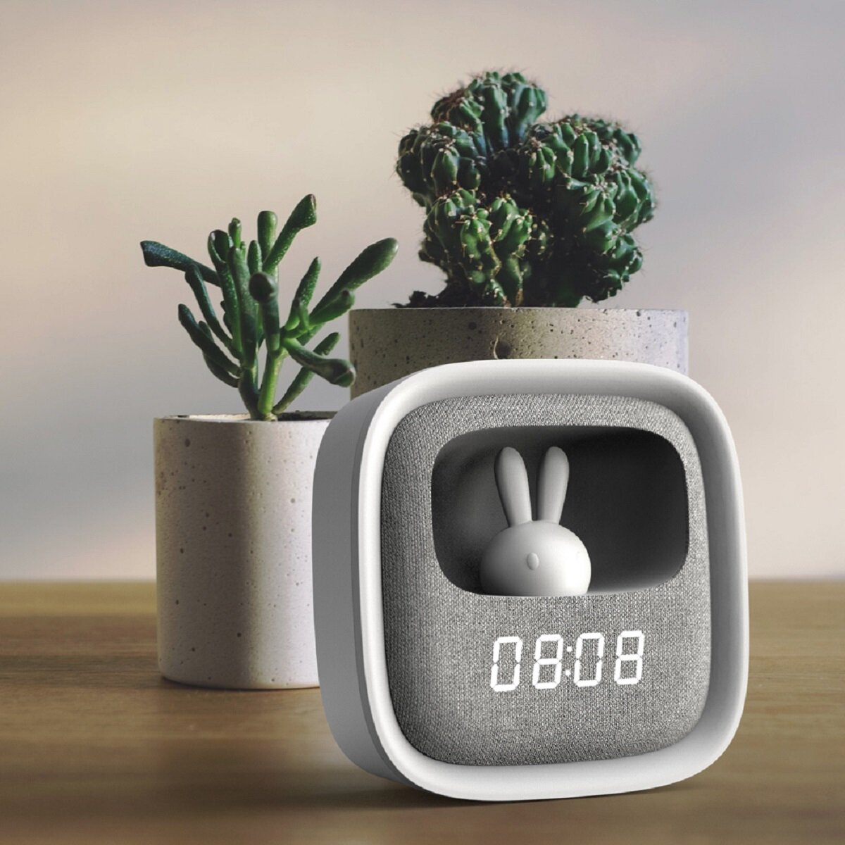 Horloge numérique blanche et grise avec une figurine de lapin à l'intérieur. Deux plantes en pot en arrière-plan.