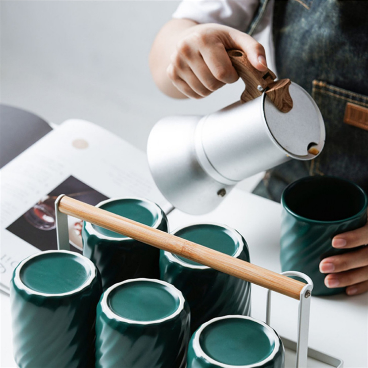 Person gießt aus einer silbernen Kaffeekanne in grüne Tassen auf einem Metallständer.