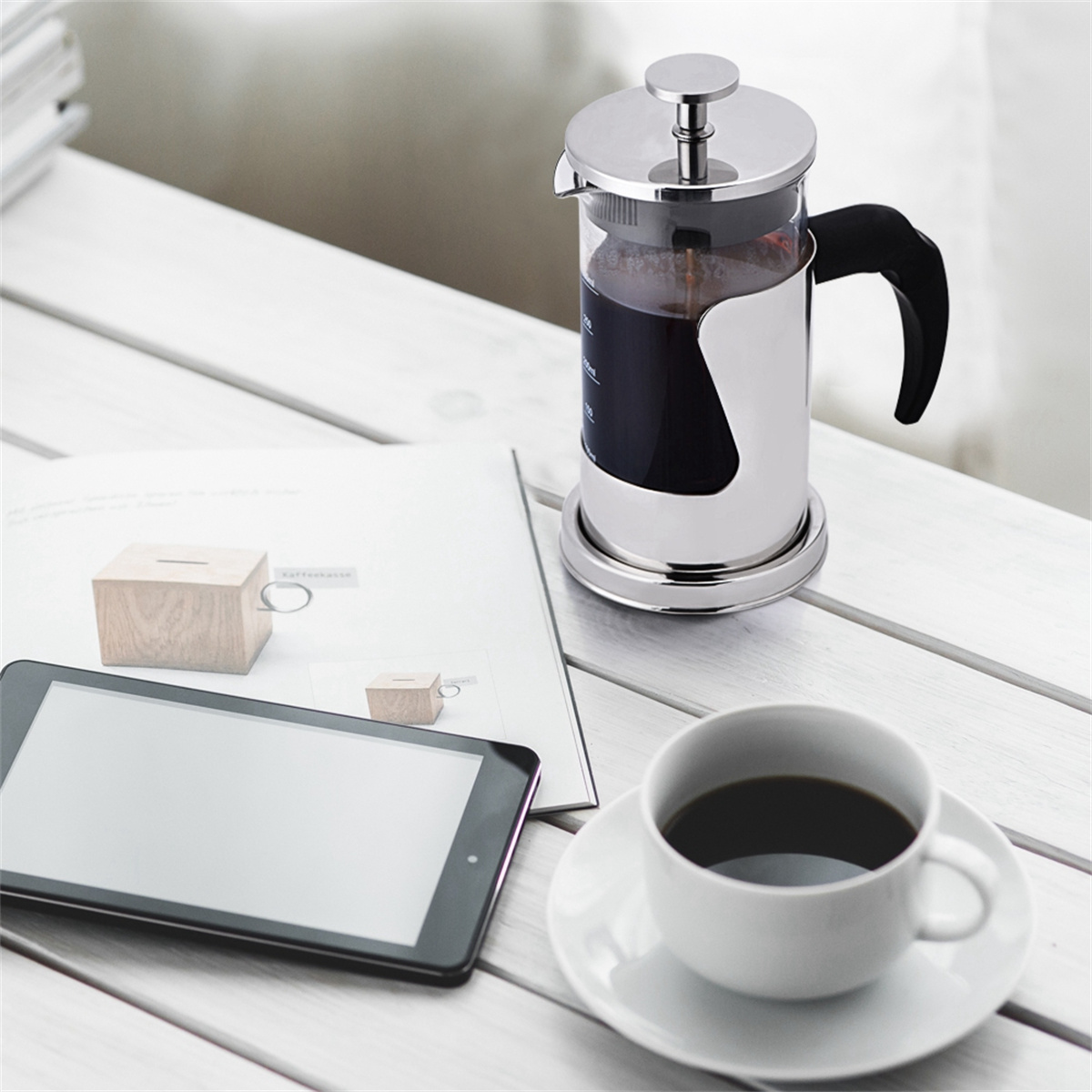 Ein Kaffee-Setup. Eine French Press, eine Tasse Kaffee, ein Tablet und eine Zeitschrift liegen auf einem weißen Holztisch.
